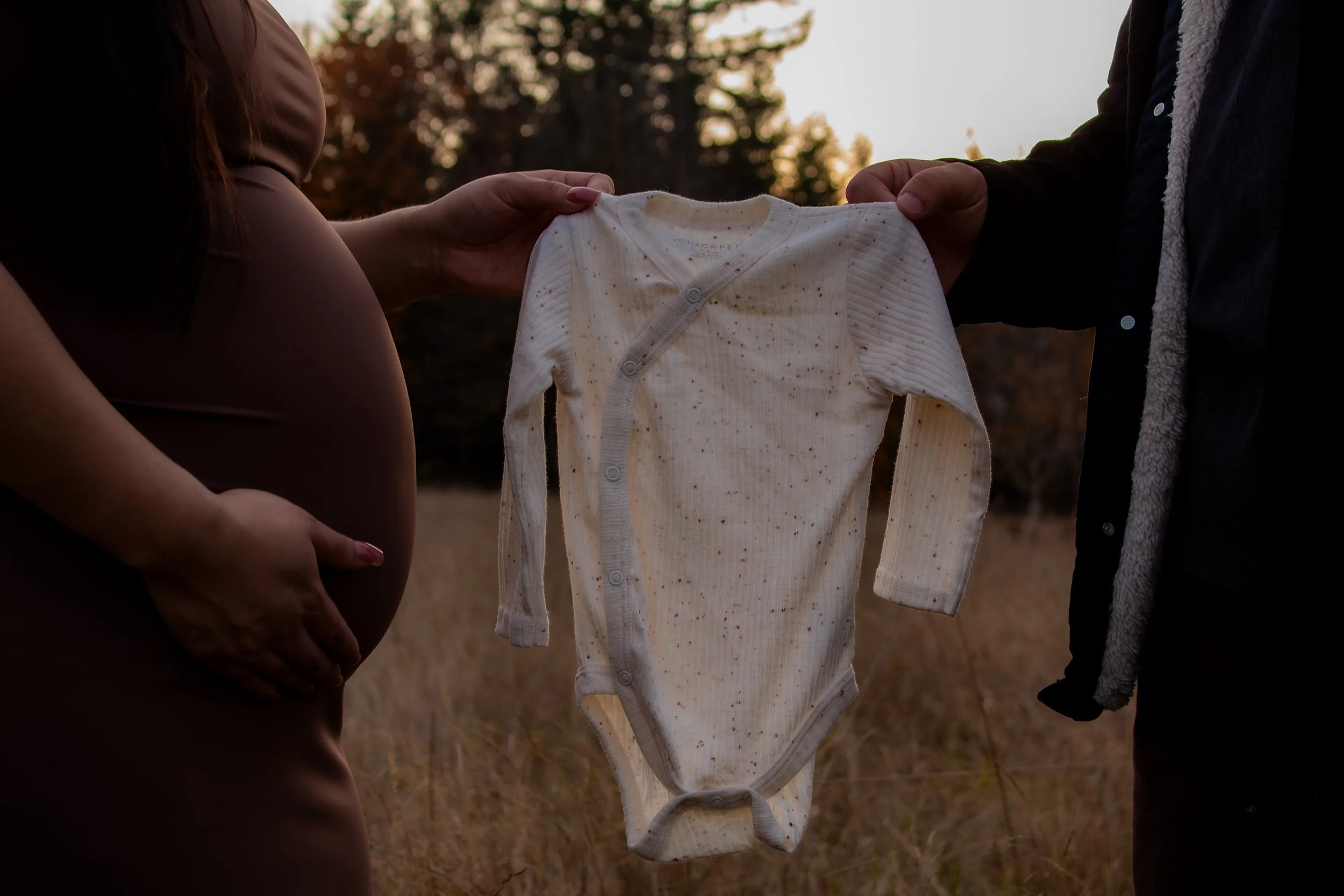 A pregnant woman and a partner holding a baby's onesie outdoors during sunset, with trees in the background.