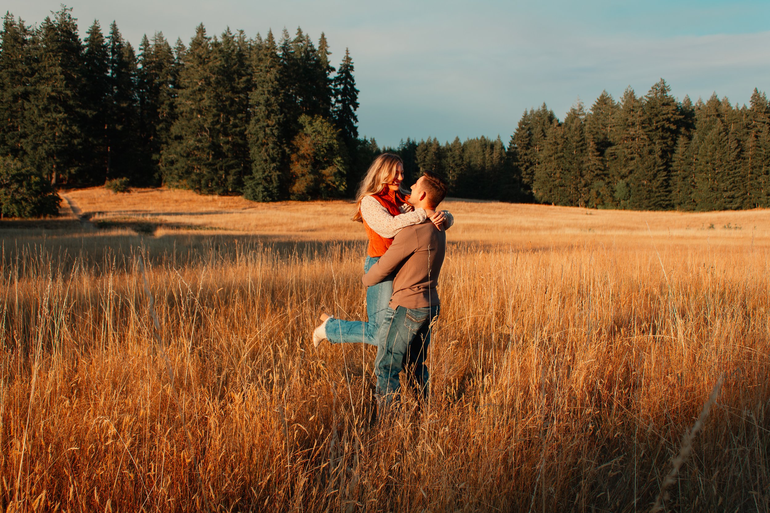 A couple embracing in a golden field with a forested background during sunset.
