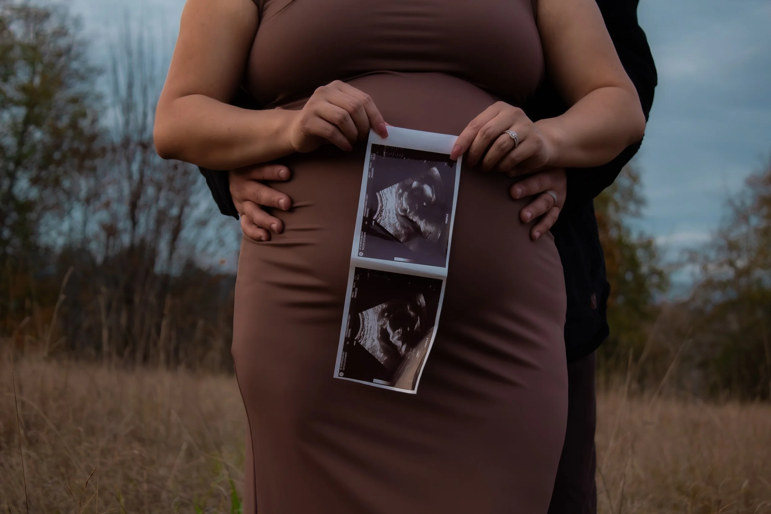 A pregnant woman holding an ultrasound image of her baby in front of her belly, with a man standing behind her and his hands on her waist, outdoors in a grassy area.
