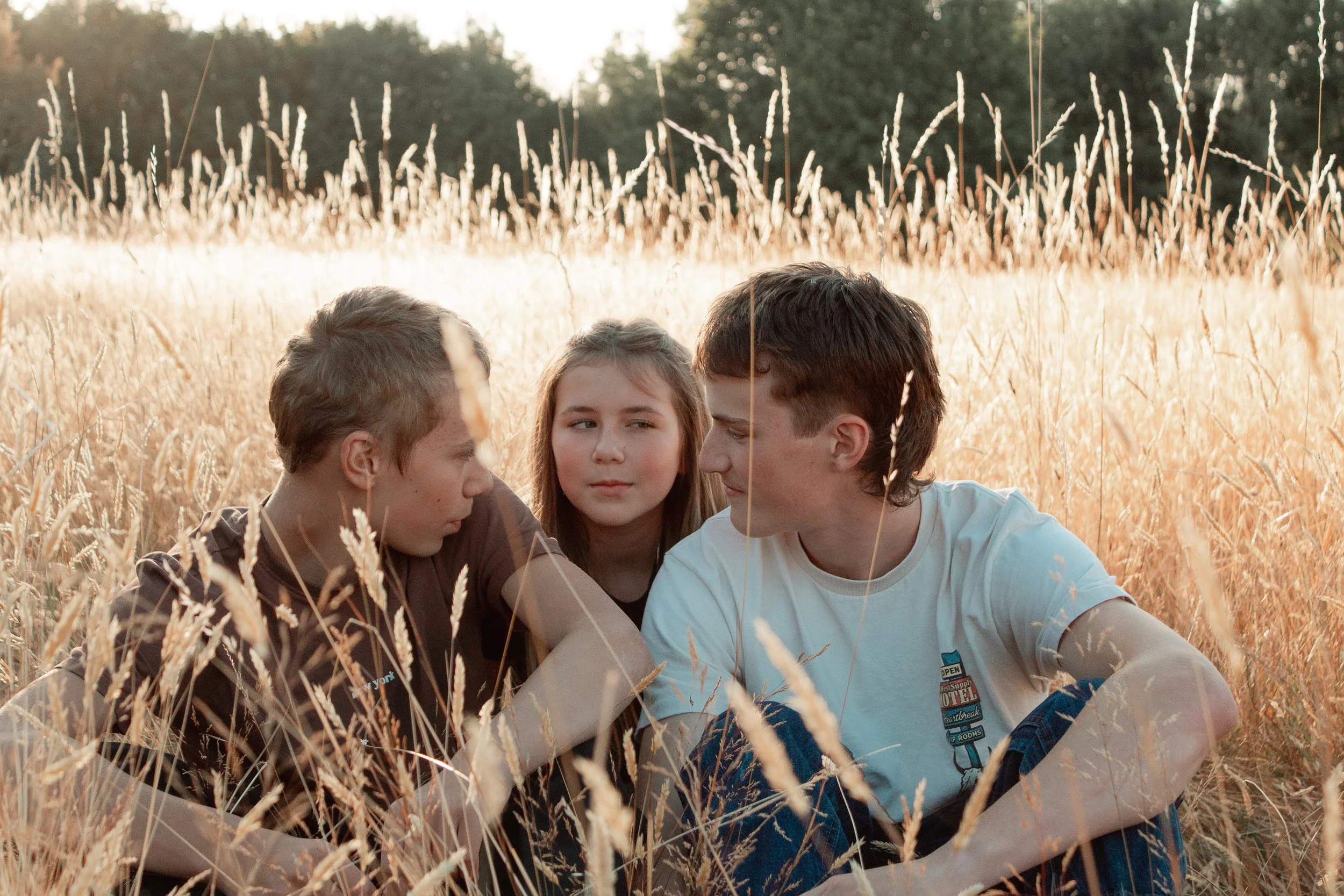 Three teenagers sitting closely in a golden wheat field during sunset, engaging in a conversation.