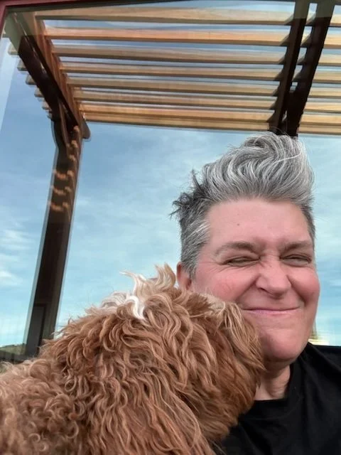 Person with short gray hair smiling and holding a curly brown dog outdoors under a pergola with a blue sky in the background.