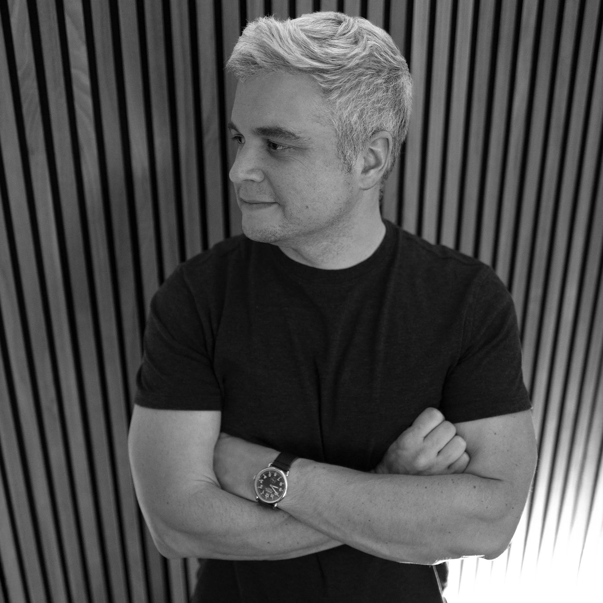 Black and white photo of a man with light-colored hair, wearing a dark T-shirt, standing with arms crossed in front of a wooden slat wall.