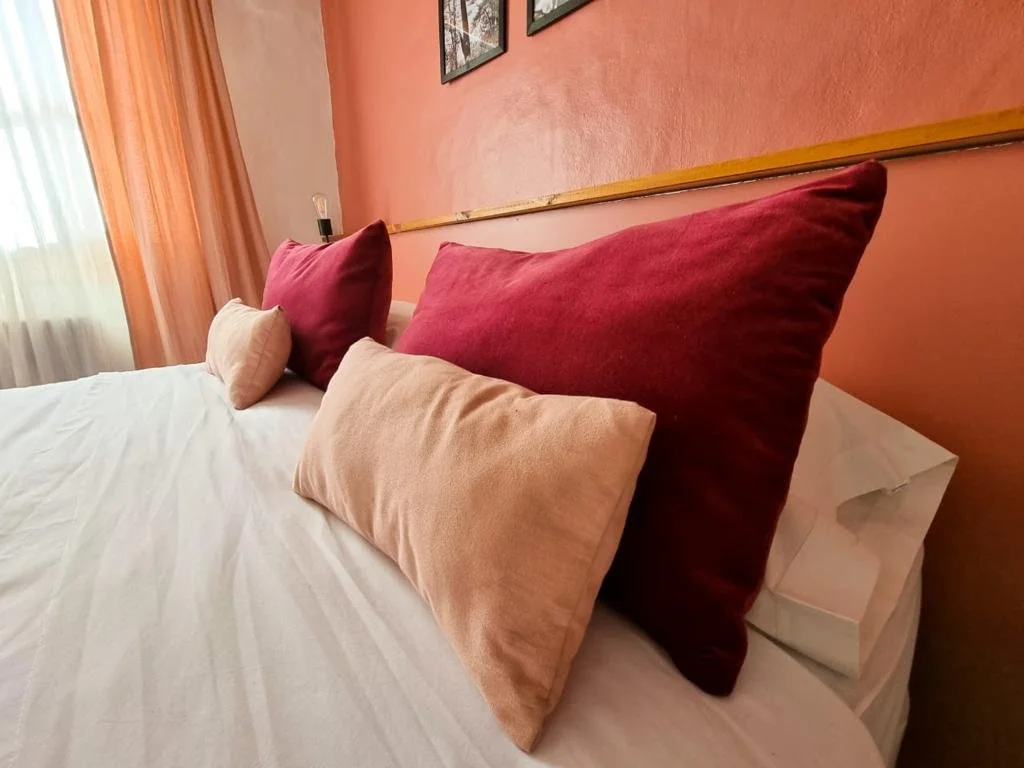 Cama con cuatro almohadas, dos grandes de color rojo y dos pequeñas de color beige, en una habitación con paredes naranjas y cortinas de color durazno.