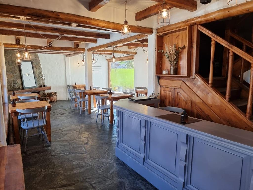 Interior de un restaurante con mesas de madera, sillas, decoración rústica, paredes de piedra y madera, luces colgantes, barra blanca y escaleras de madera.