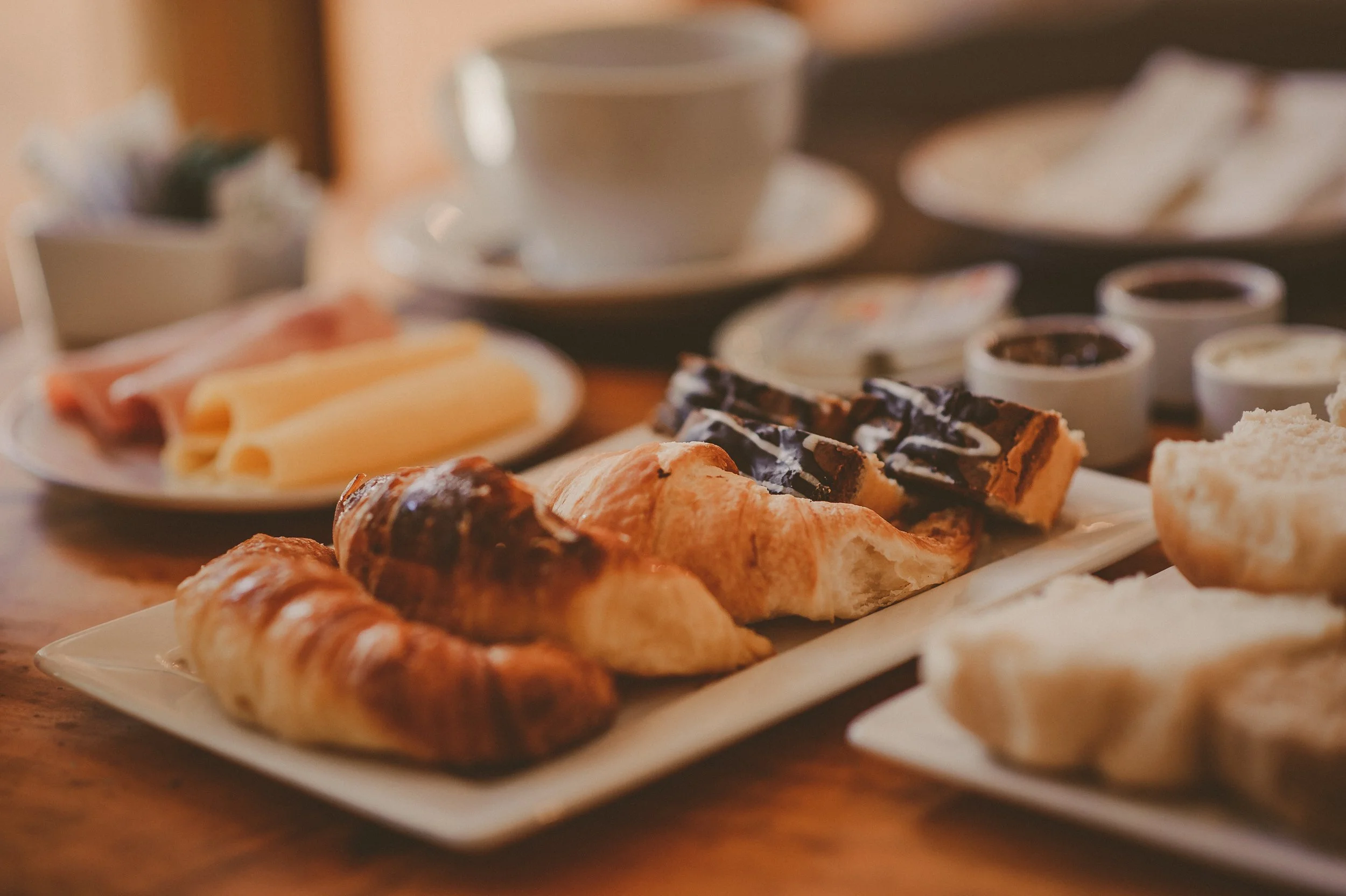 Variados alimentos y postres en bandejas y platillos, incluyendo croissants, pasteles, galletas, y pequeños recipientes con salsas o dips, en un ambiente de desayuno o merienda.