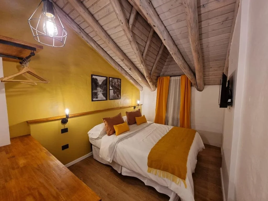 Habitación con cama, paredes blancas y amarillas, lámparas de lectura, cuadros, cortinas amarillas y naranja, techo de madera con vigas, mesas de noche y televisión.