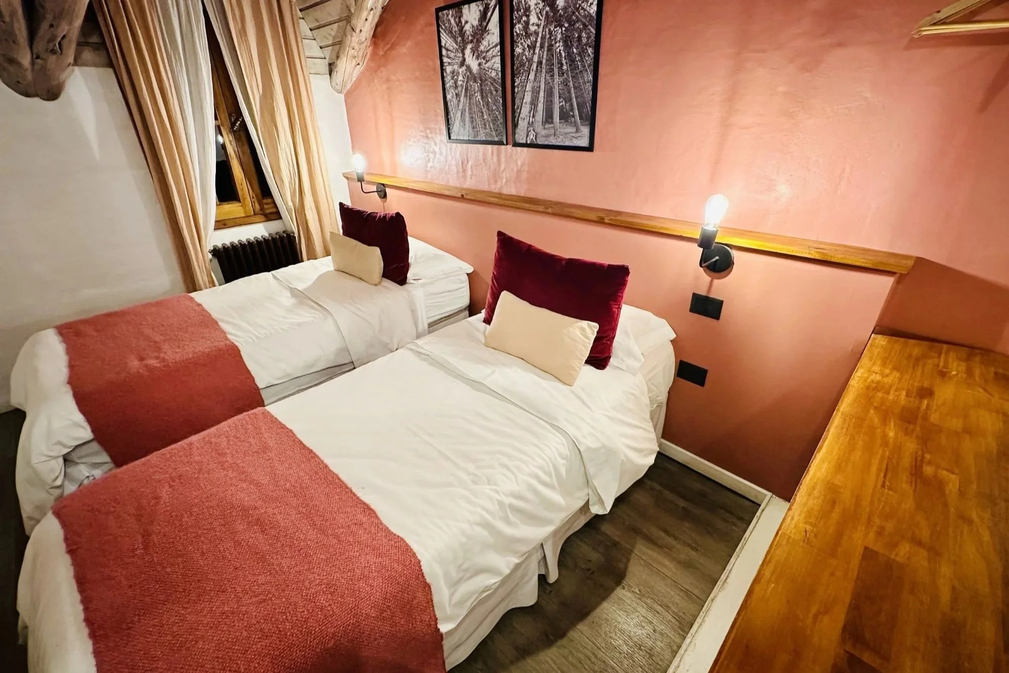 Habitación con paredes de color rosa, dos camas con sábanas blancas y almohadas beige y burdeos, cortinas beige en una ventana de madera, dos cuadros con escenas en blanco y negro en la pared y lámparas de lectura en la cabecera de cada cama, mesa de madera al lado.