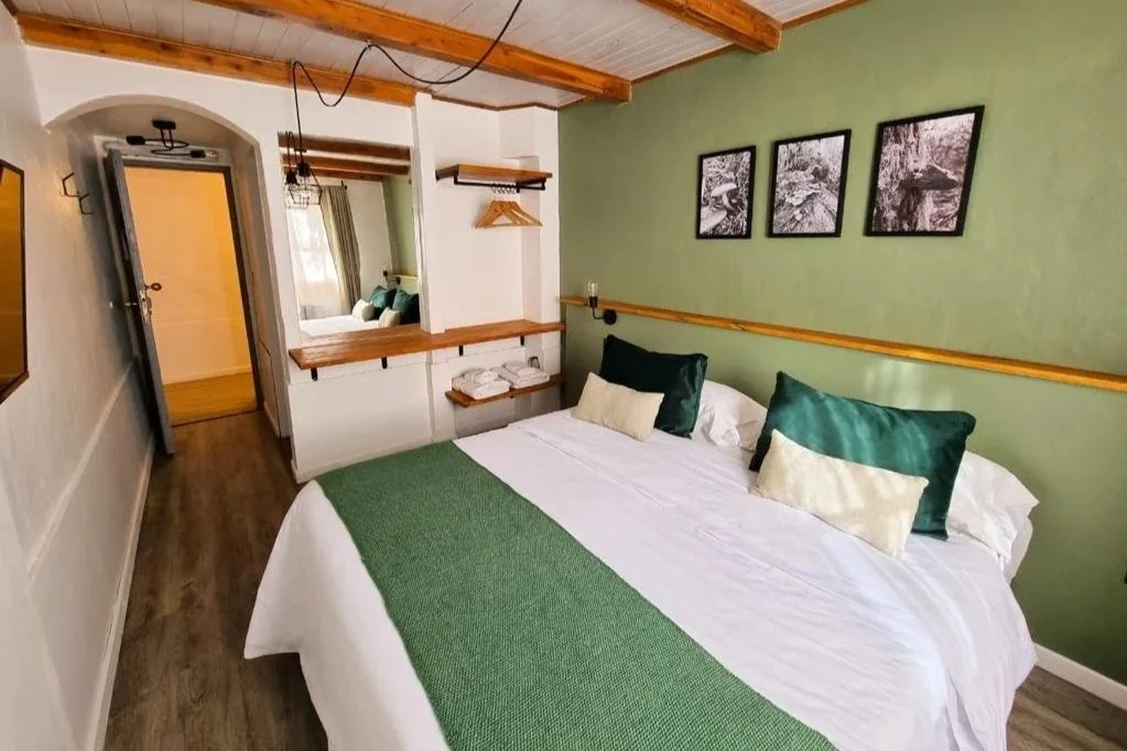 Habitación con cama grande, almohadas verdes y blancas, paredes verdes, cuadros en la pared y área de descanso separada con espejo y ventanas.