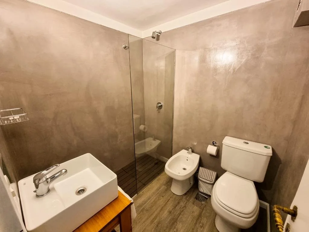 Baño pequeño con lavabo, inodoro, bidé y ducha con cancel de cristal, paredes de color beige y suelo de madera.