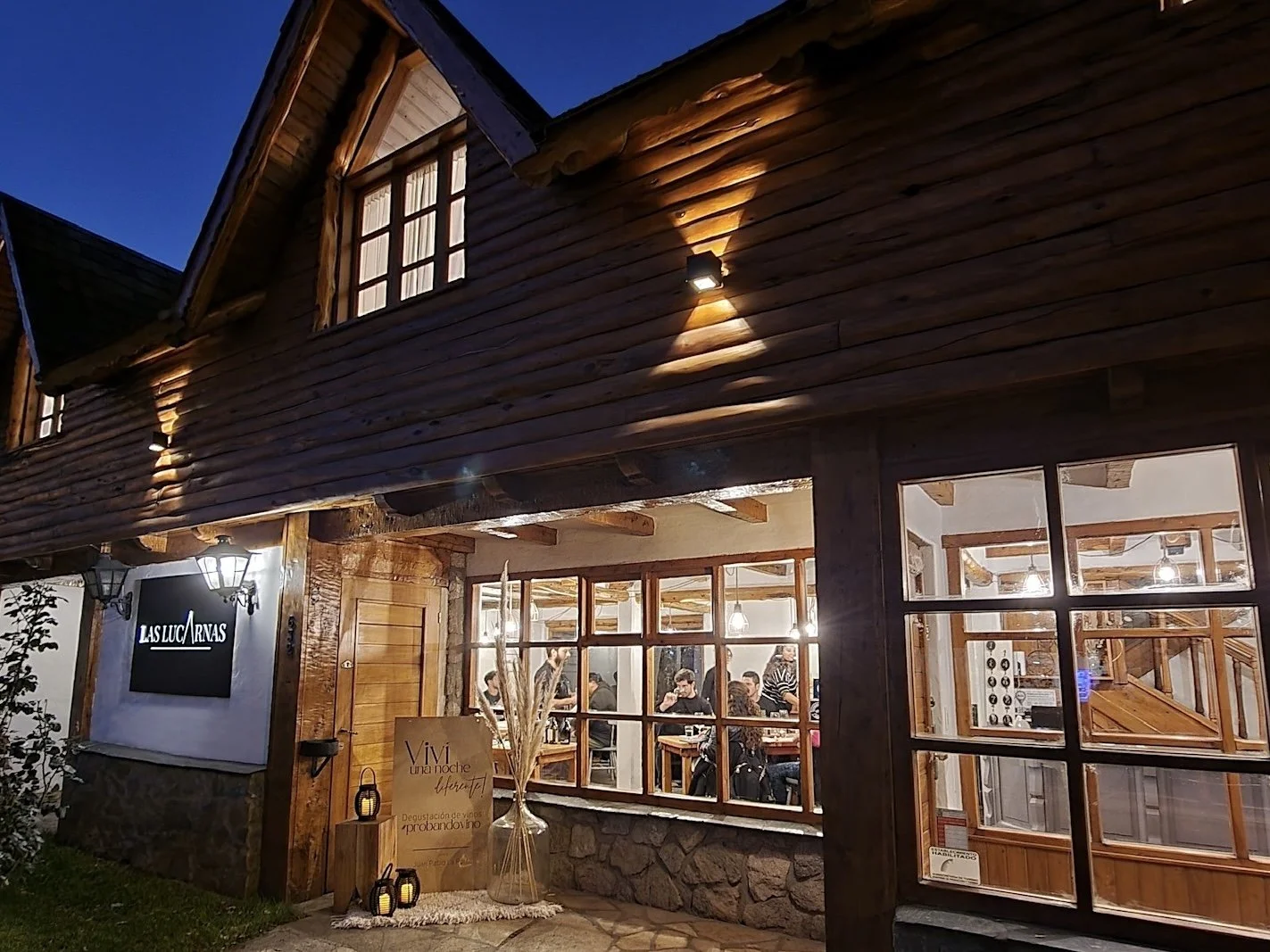 Vista nocturna de una casa de madera con ventanas grandes, mostrando un interior con varias personas sentadas y conversando, luz cálida y ambiente acogedor.