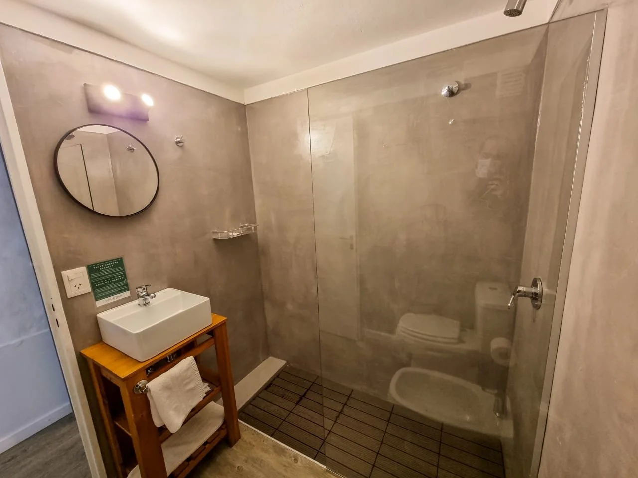 Baño con área de ducha de vidrio, lavabo sobre mueble de madera, espejo redondo, y sanitario visible reflejado en la ducha.