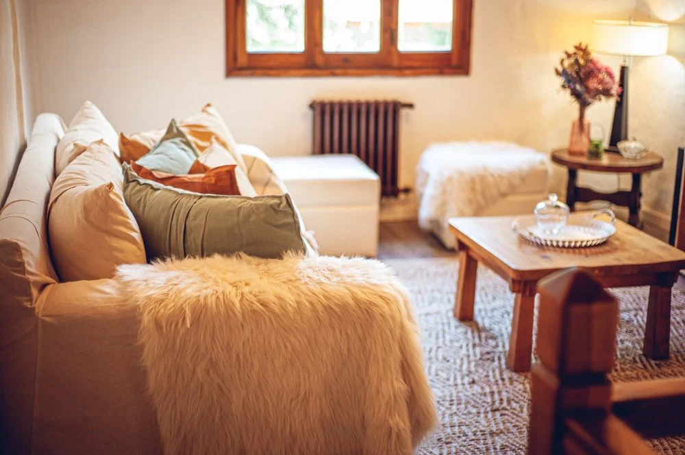 Sala de estar con sillón beige con muchos cojines, un sofá con mantas y una mesa de centro de madera con una bandeja y decór, ventanas con vista a la naturaleza y una lámpara de pie con flores.