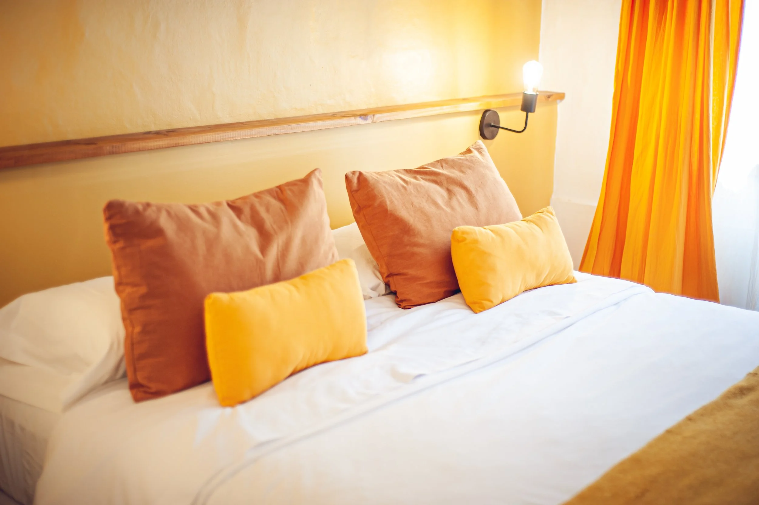 Cama con varias almohadas en tonos cálidos y cortinas amarillas y anaranjadas, lámpara de pared encendida en habitación.