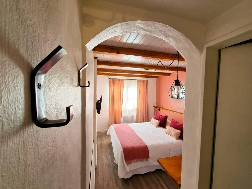 Vista de una habitación de hotel con cama, cortinas color durazno, pared rosa, lámpara decorativa y muebles de madera.