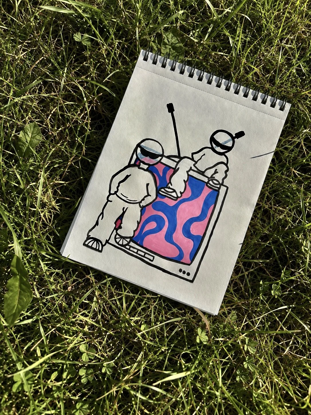 Dessin dans un carnet posé sur l’herbe, illustrant deux figures portant des vêtements amples, adossé et assis à une télévision diffusant des formes abstraites rose et bleu. 