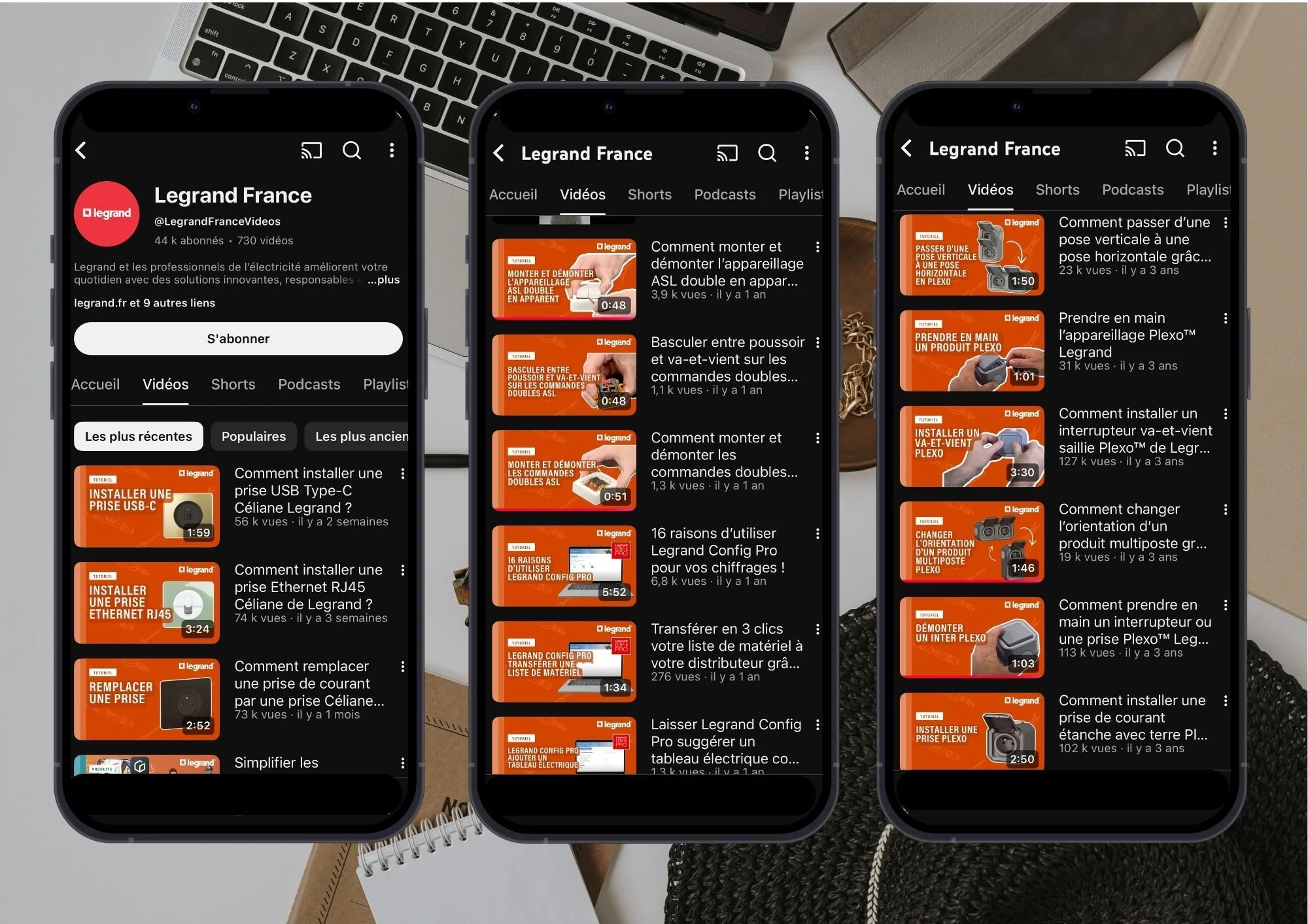 Trois téléphones portables affichant la chaîne YouTube Legrand France, avec des vidéos sur l'installation électrique, le branchement et la configuration d'appareils électriques, placés sur un bureau avec un clavier, un cahier et des objets décoratifs en arrière-plan.