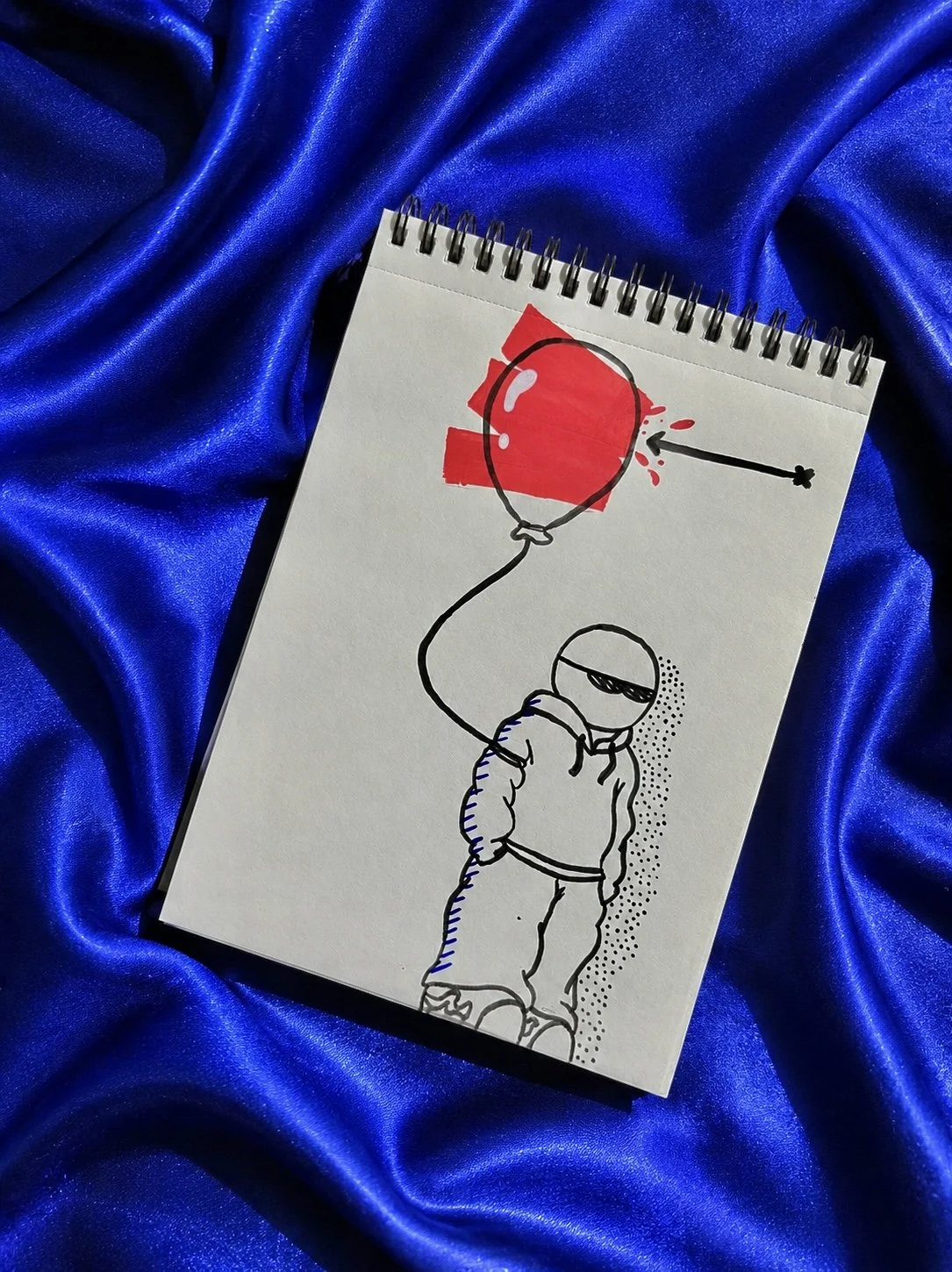 Dessin dans un carnet posé sur un tissu de soie Bleau vif, illustrant une figure portant un sweat à capuche, tenant un ballon rouge. 