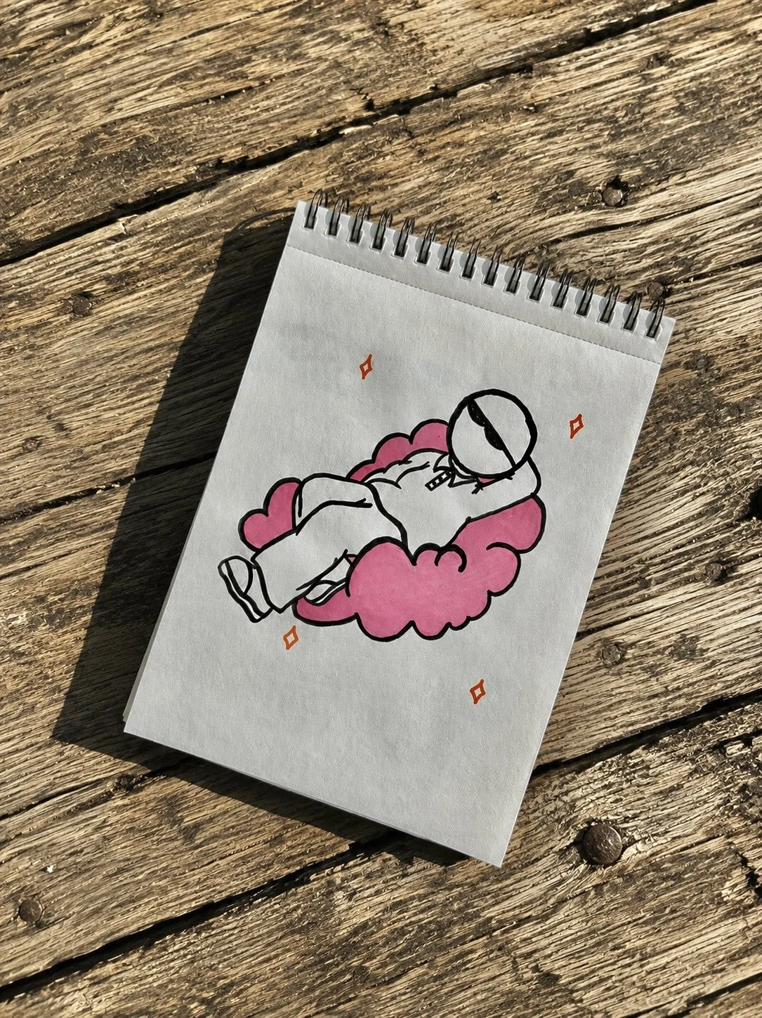 Dessin d'un personnage, allongé sur un nuage rose, sur une feuille de papier posée sur une surface en bois.