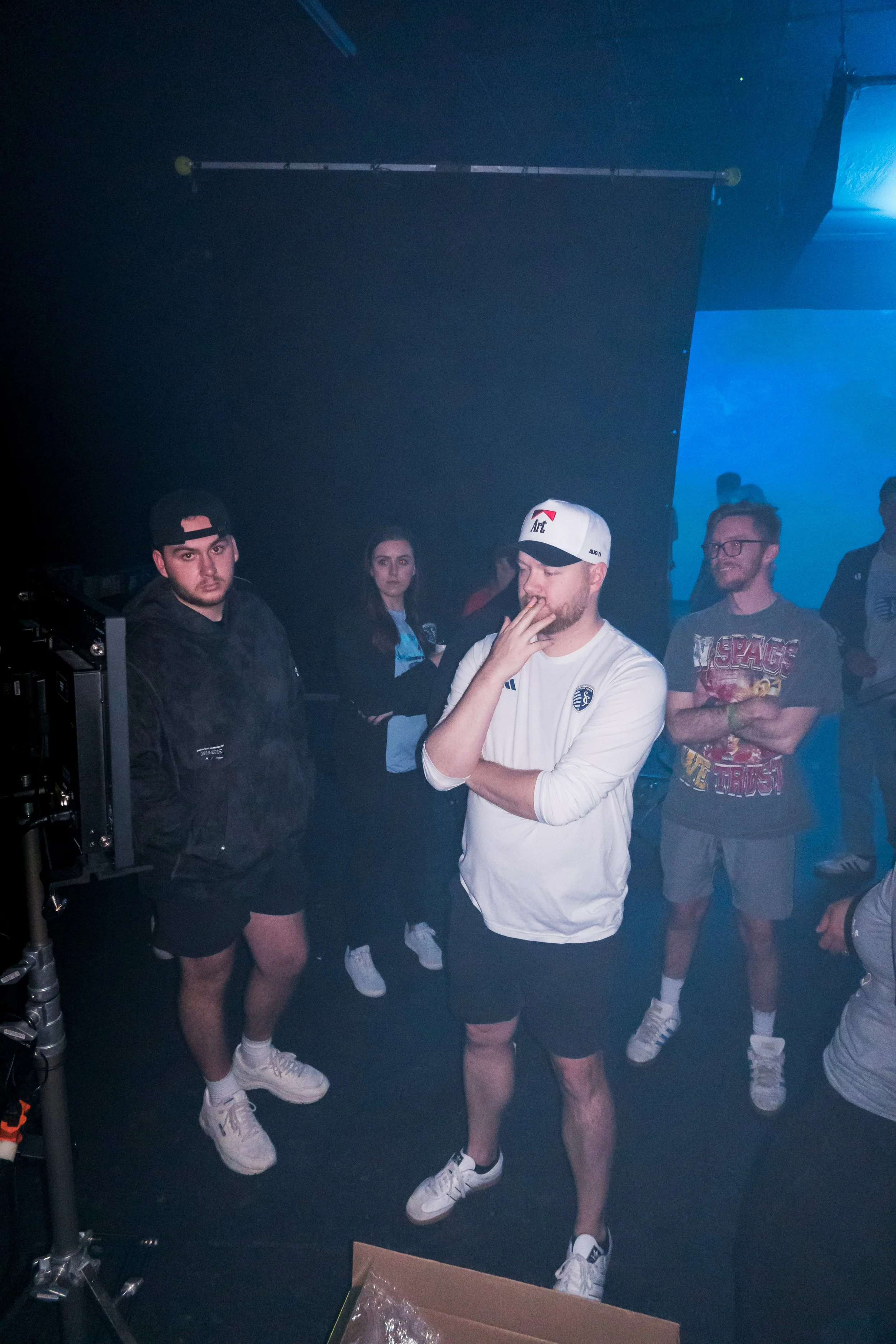 Group of people at a production shoot, one man in the center with hand over mouth, wearing a white shirt and cap, others standing around in a dimly lit setting.