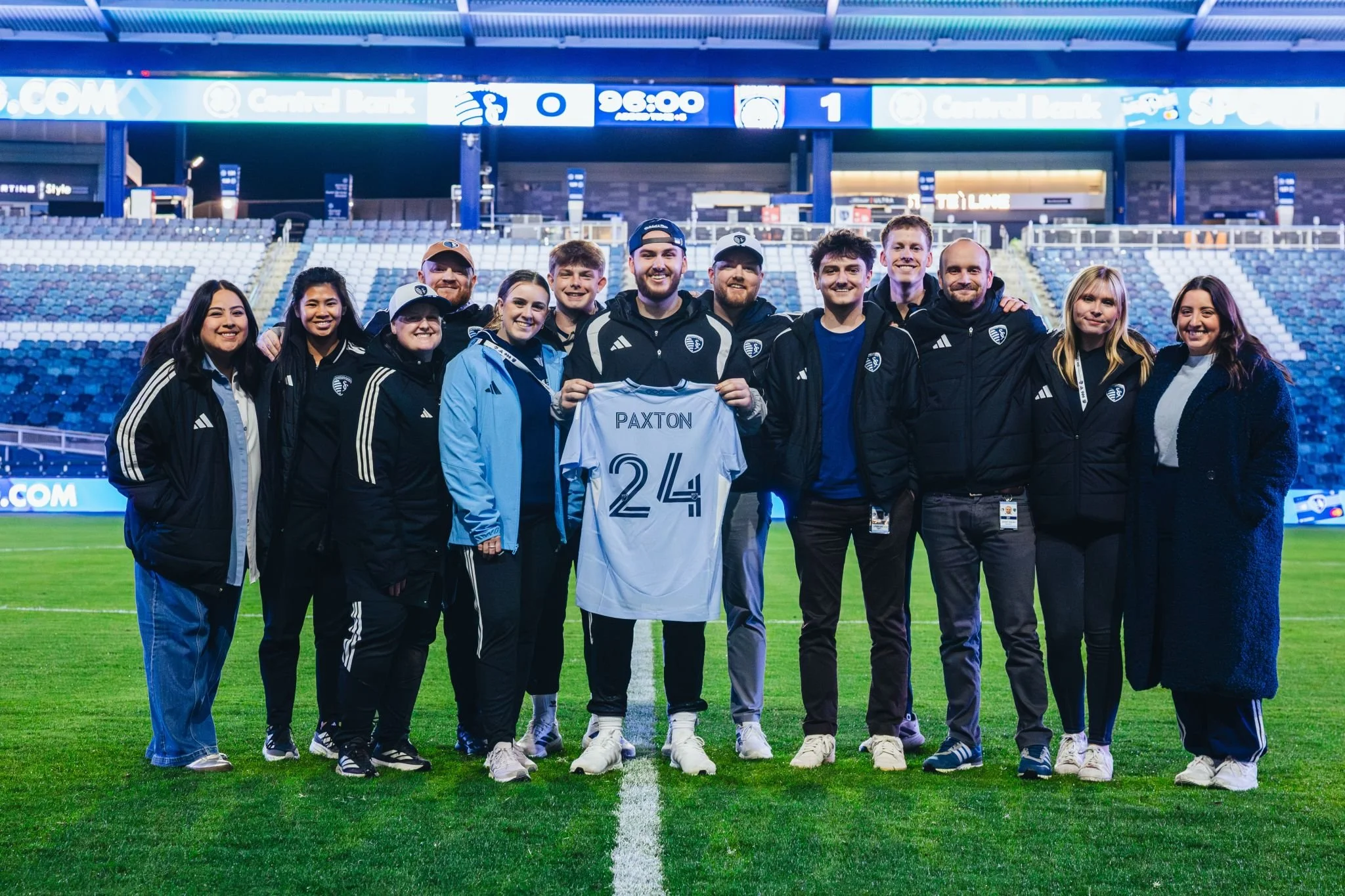 Group of soccer team and staff standing on a field at a stadium, holding a jersey with the name 'PAXTON' and the number 24, smiling for a photo.
