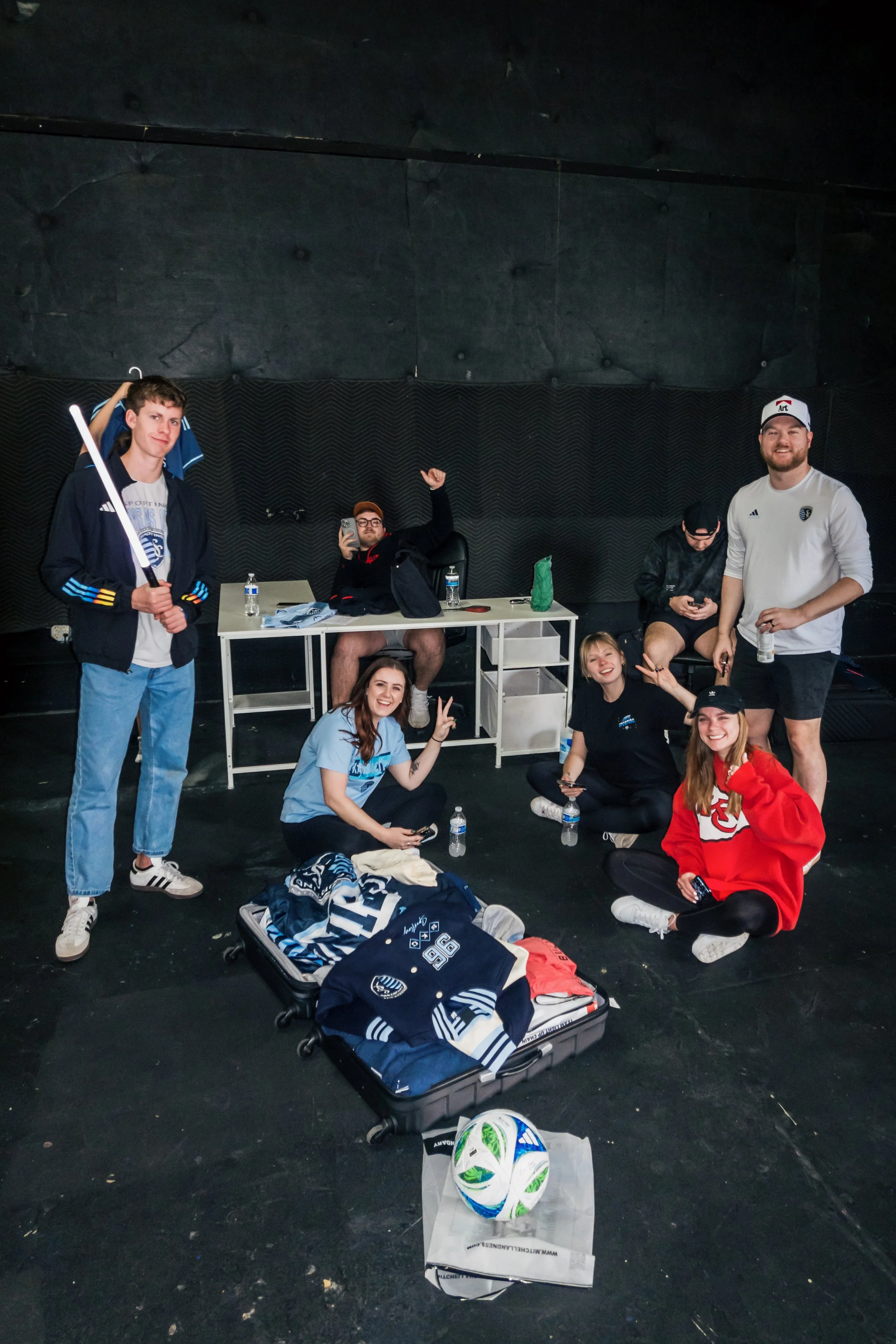 A group of seven young adults with sports gear and water bottles in a room with a black wall, sitting and standing around an open suitcase, with a soccer ball on the floor.