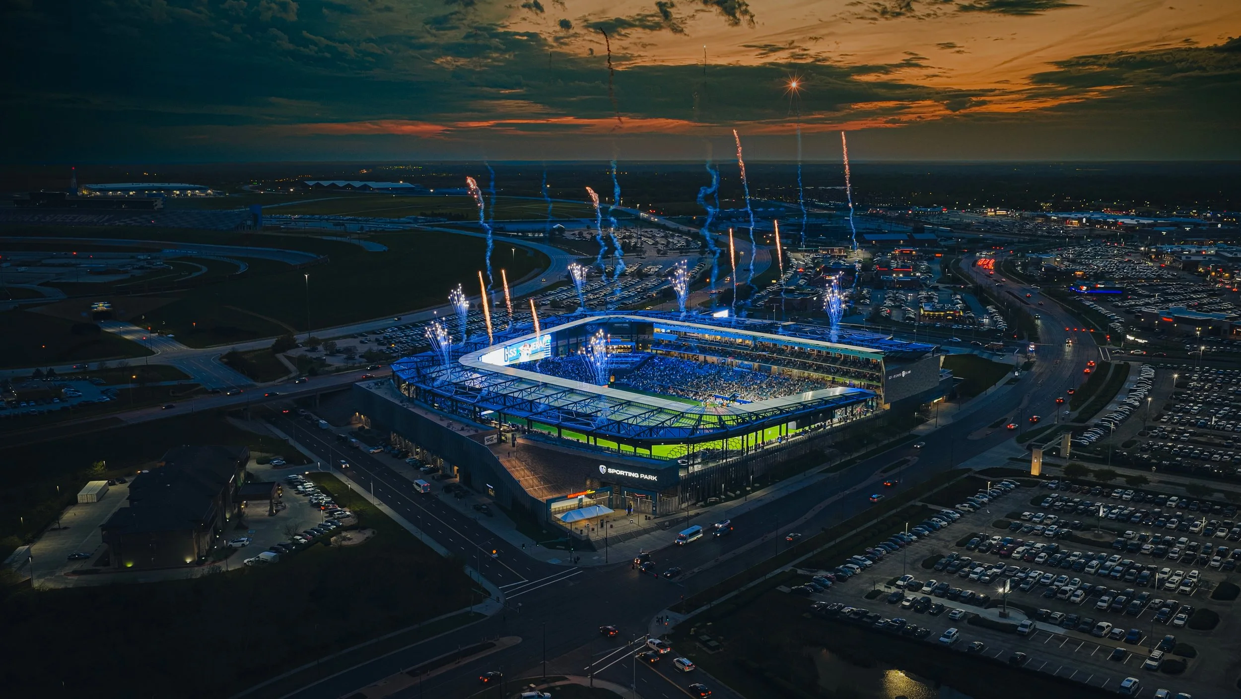 An aerial night view of Sporting Park with fireworks and a bright display, surrounded by parking lots and roads, with a sunset sky in the background.
