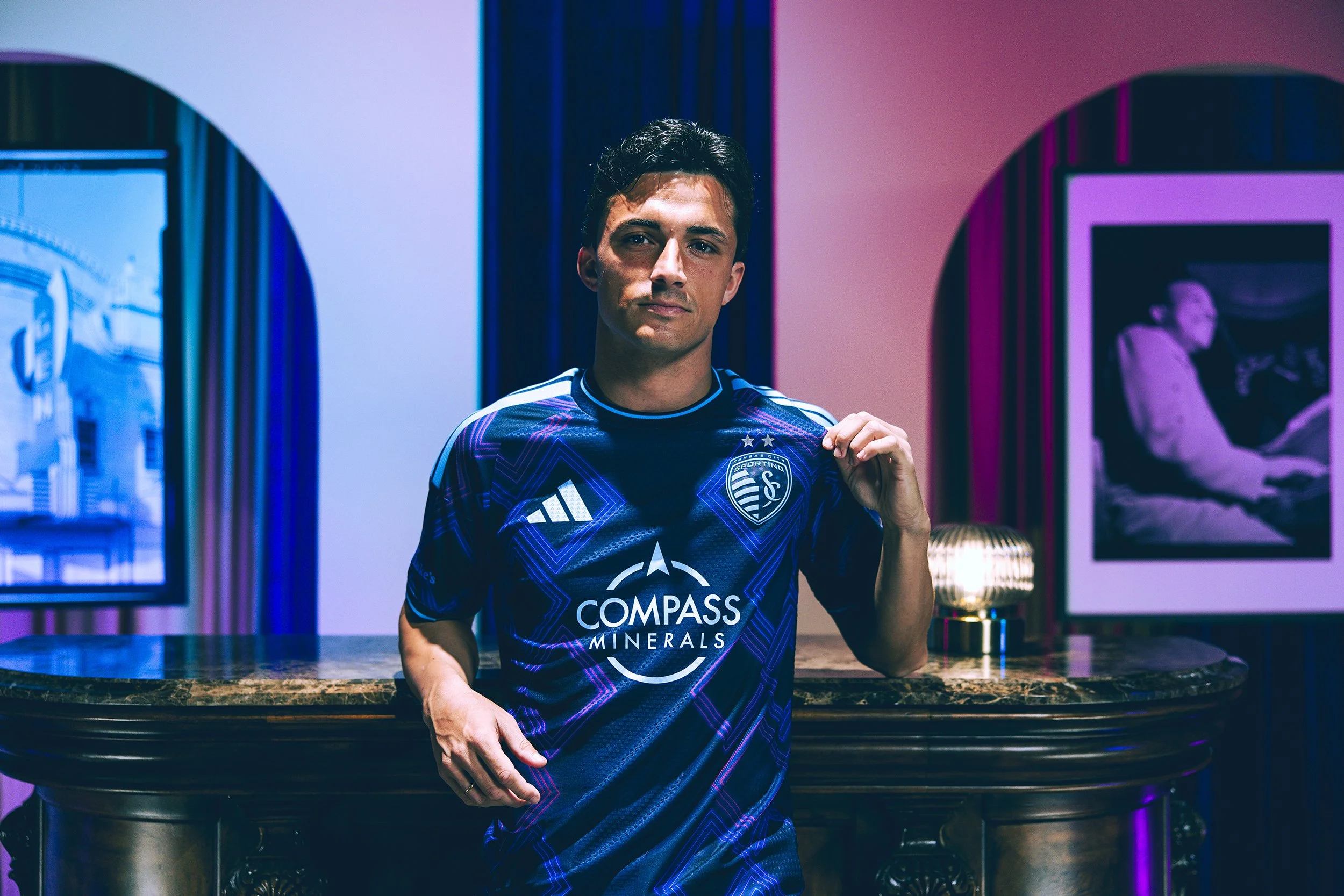 A man wearing a blue soccer jersey with sponsorship logos standing at a bar in a dimly lit room.