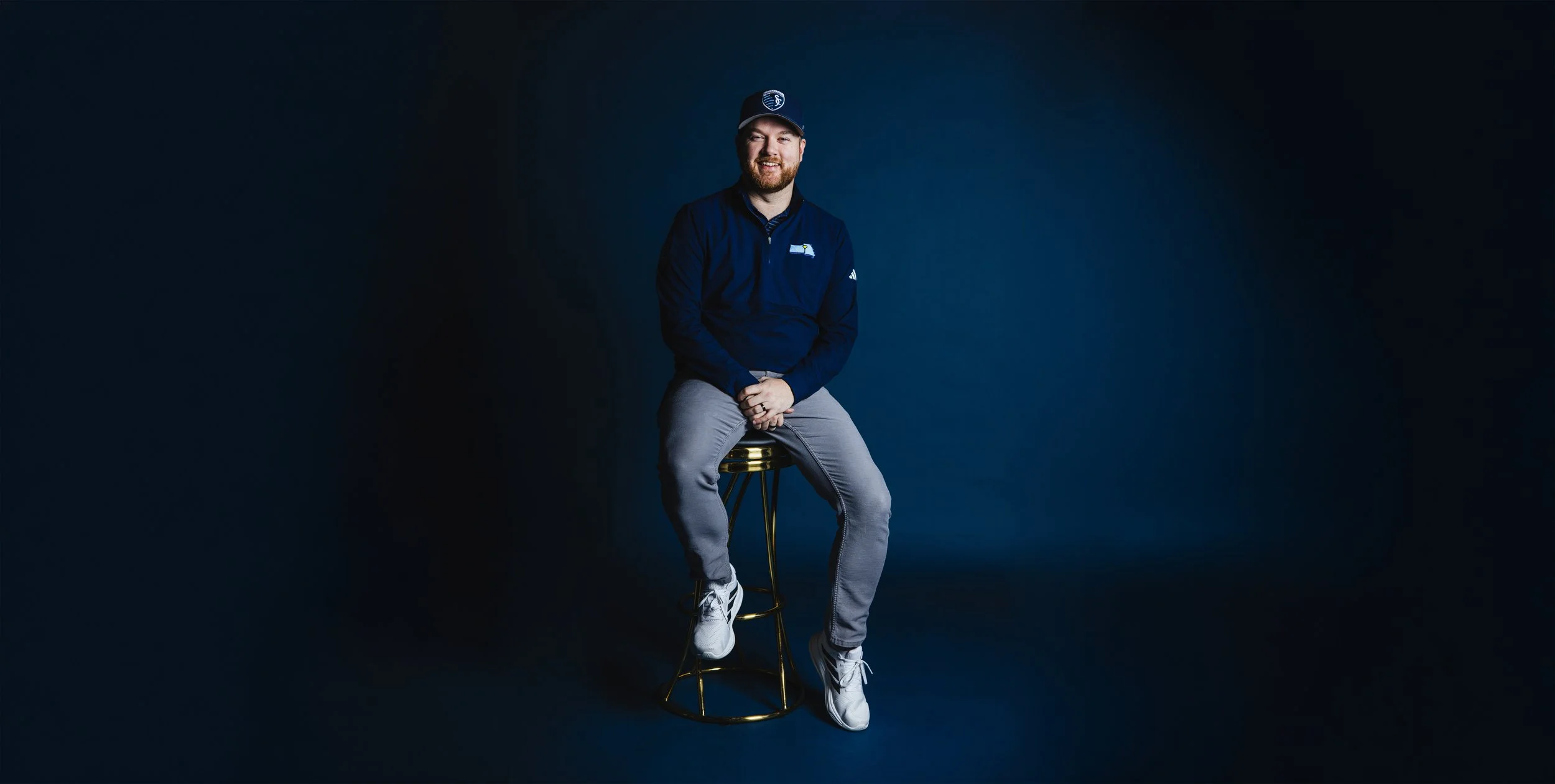 A man sitting on a gold barstool with a dark blue background, wearing a navy blue jacket, a cap, gray pants, and white sneakers, smiling.