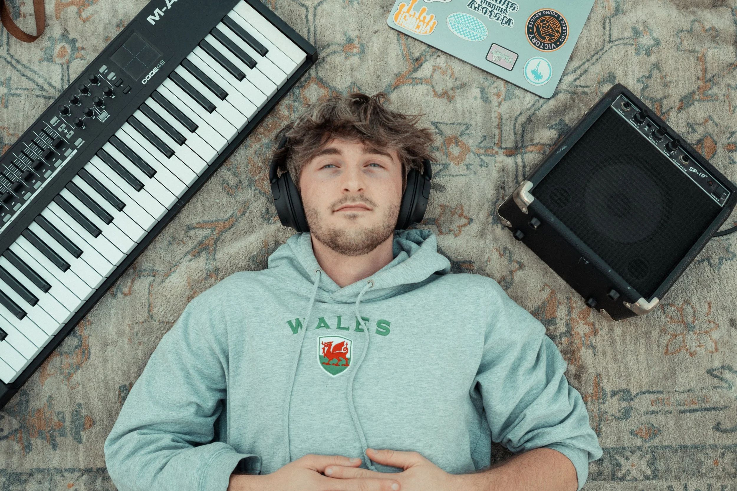 A young man with headphones lying on a patterned rug with a keyboard and an amplifier around him.