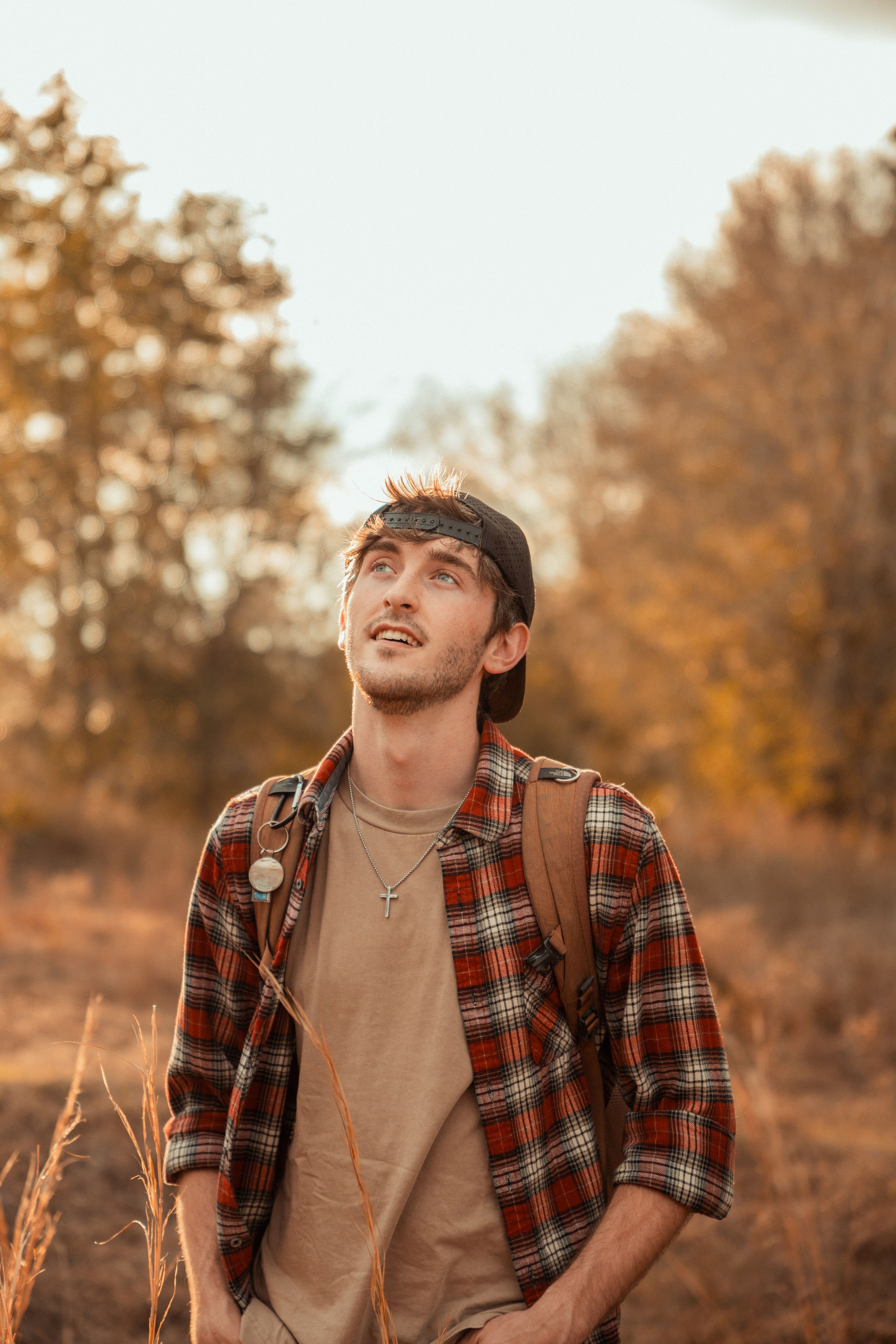 A young man with a beard and wearing a black baseball cap backwards, a red plaid shirt, and a beige t-shirt, standing outdoors during autumn, looking up with a thoughtful expression, surrounded by trees with fall foliage.