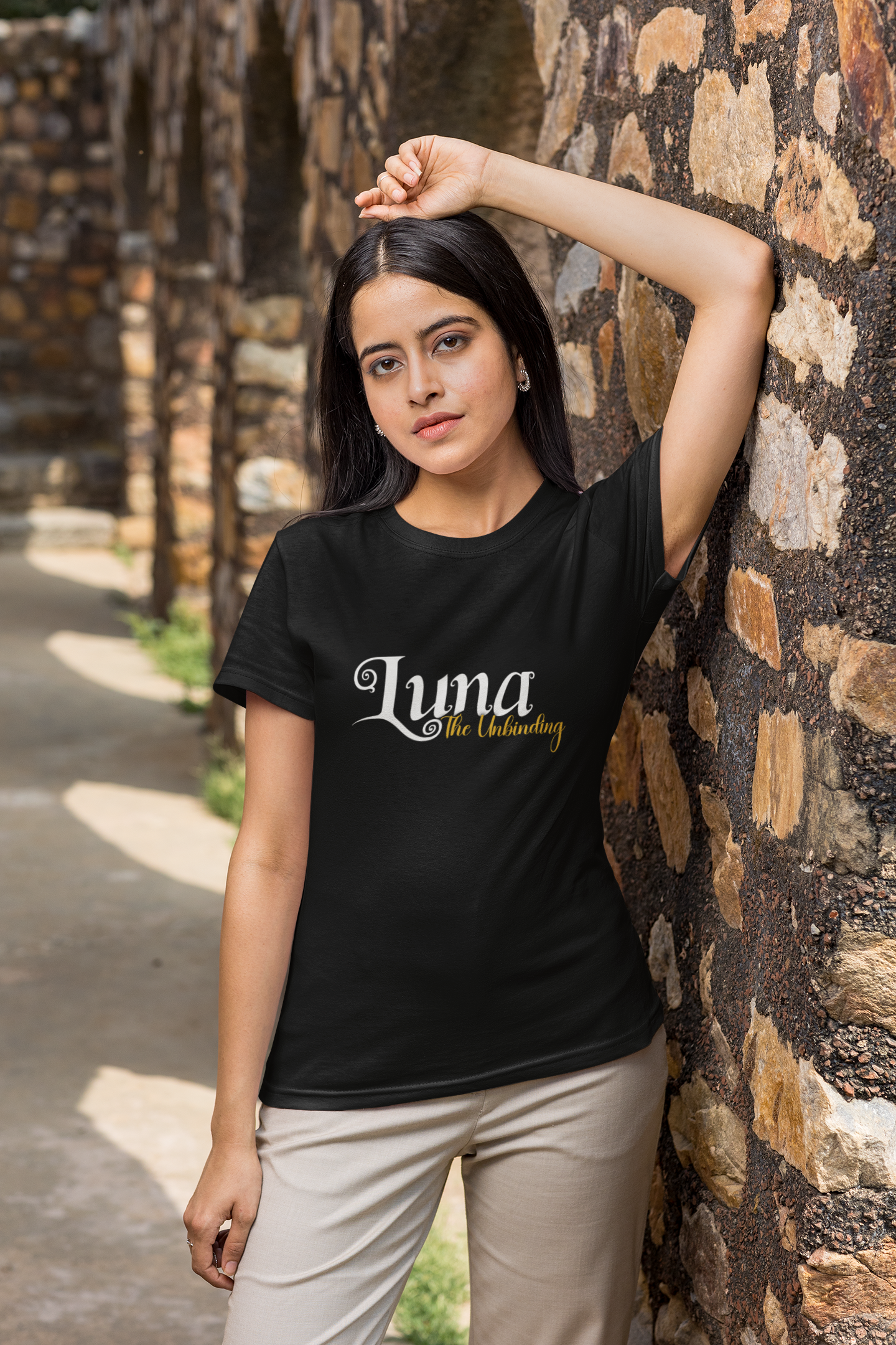 A young woman with dark hair, wearing a black t-shirt with 'Luna The Unbinding' written on it, standing outdoors against a stone wall.