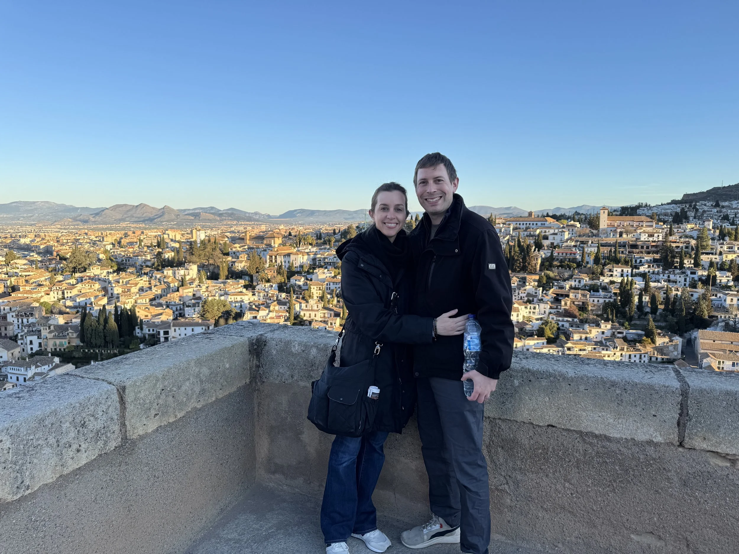An adult man and woman in their mid 30s smile for the camera while standing on the balcony of a stone building overlooking a scene of historic houses in Southern Spain.