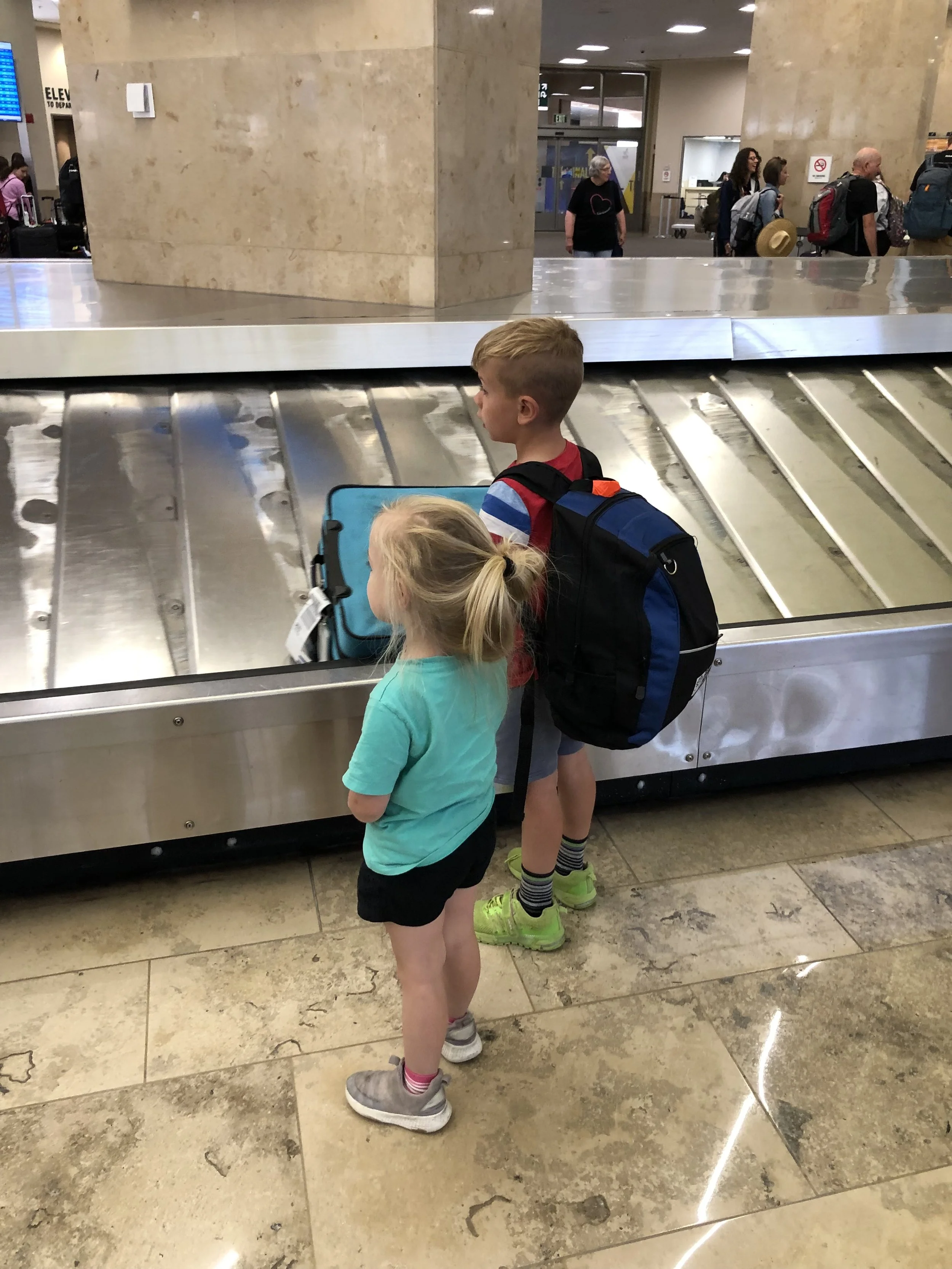 Two small children, a boy and a girl, waiting patiently to gather their luggage by an airport conveyor belt.