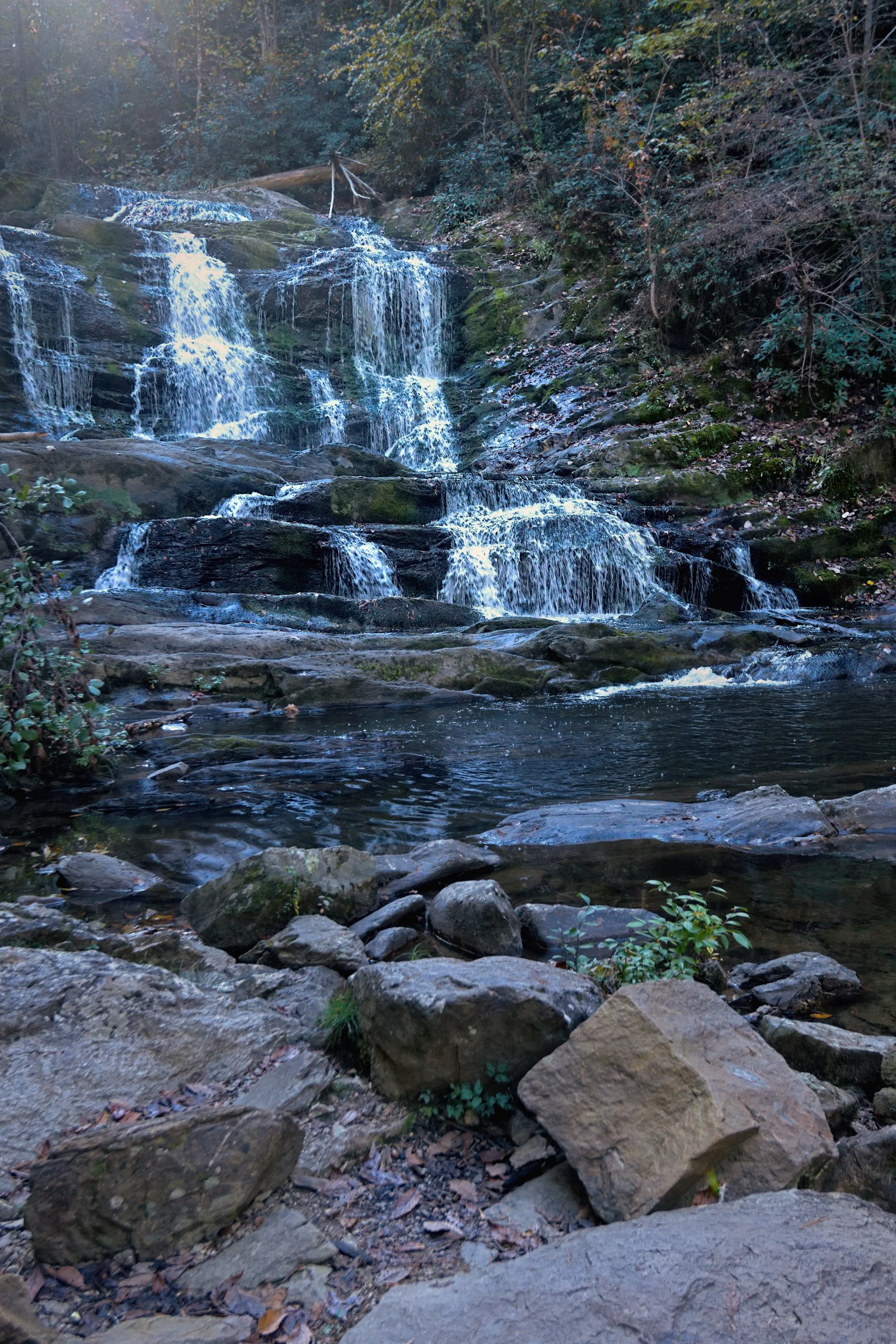 Tiered Appalachian waterfall over mossy rock ledges in soft late-season light, with cool blue water, muted greens, gray stone, and a calm reflective pool in the foreground.