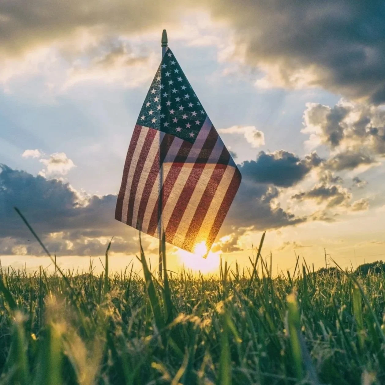 American flag planted in a grassy field at sunset with a cloudy sky.