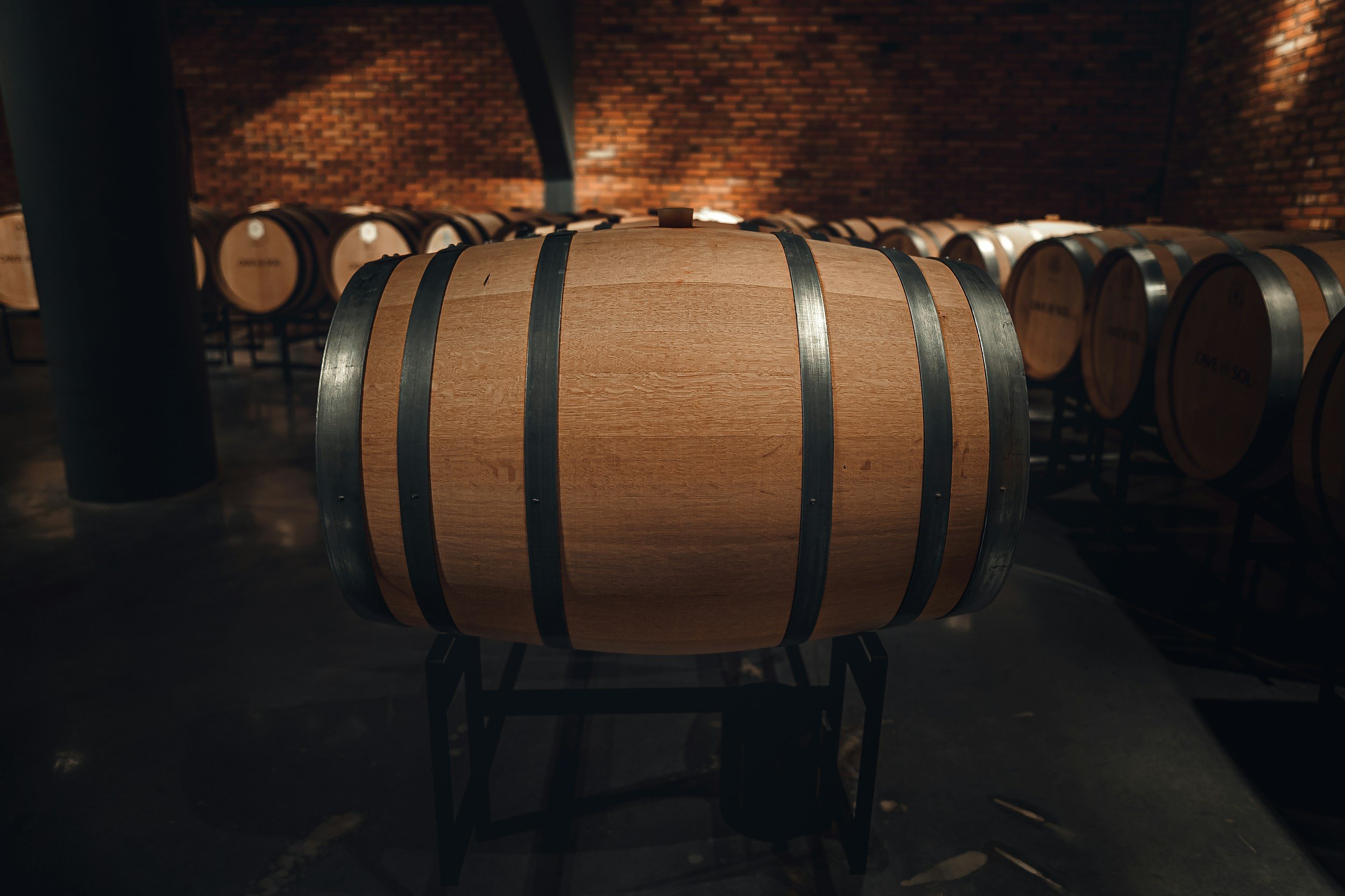 A wooden barrel with metal hoops in a wine cellar with brick walls and multiple barrels in the background.