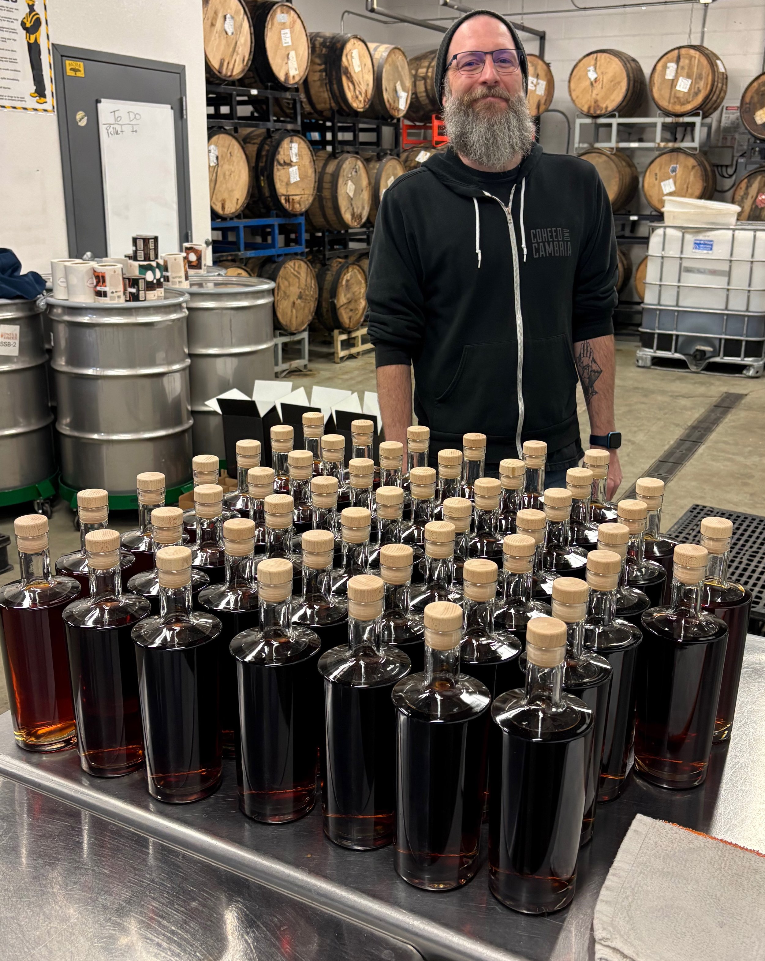 A man with a beard and glasses standing behind rows of glass bottles filled with dark liquid, in an industrial setting with barrels on racks in the background.