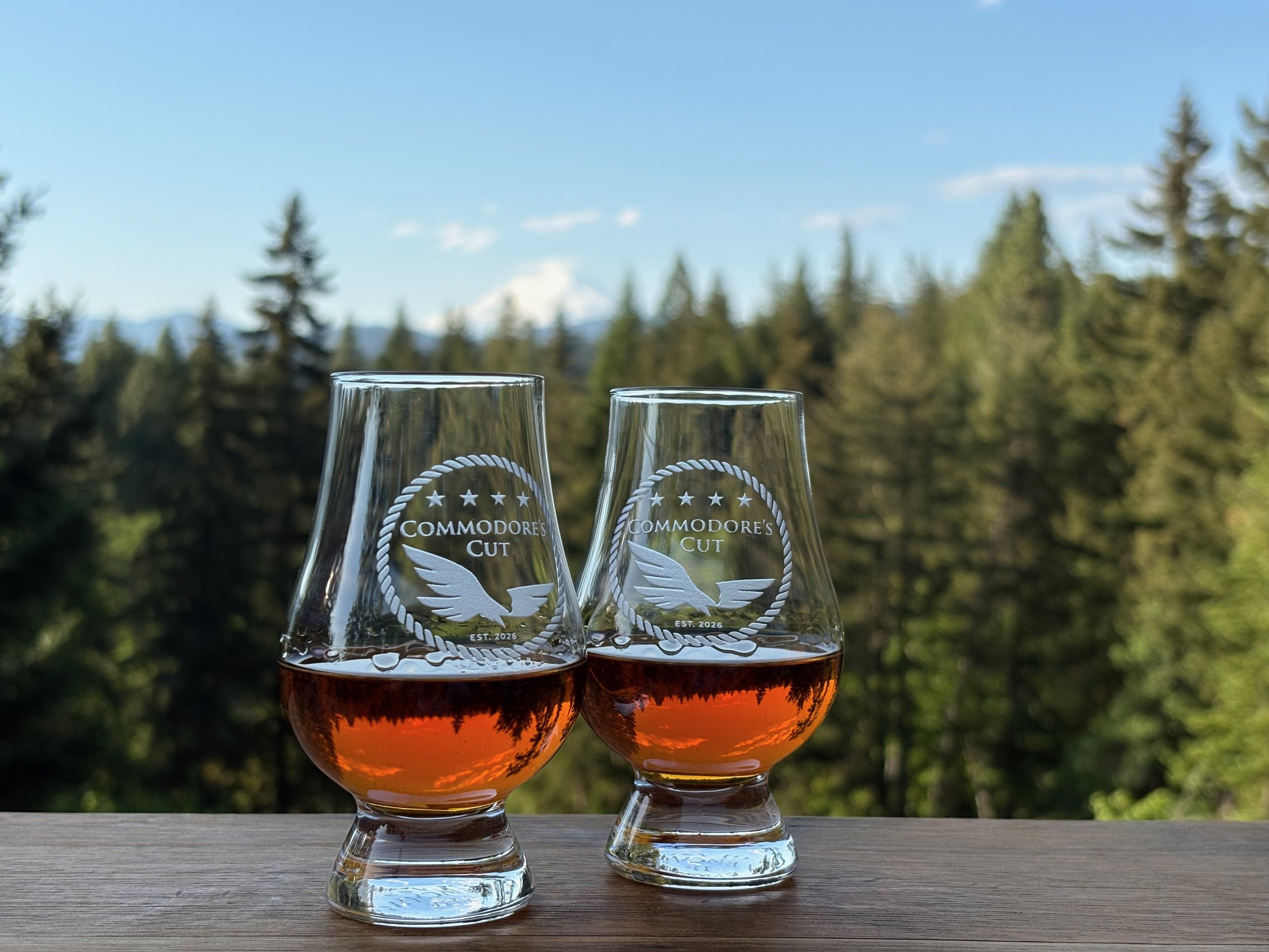 Two glasses of amber-colored beverage, etched with the logo "Commodore's Cut" , set outdoors with a background of trees and a blue sky.