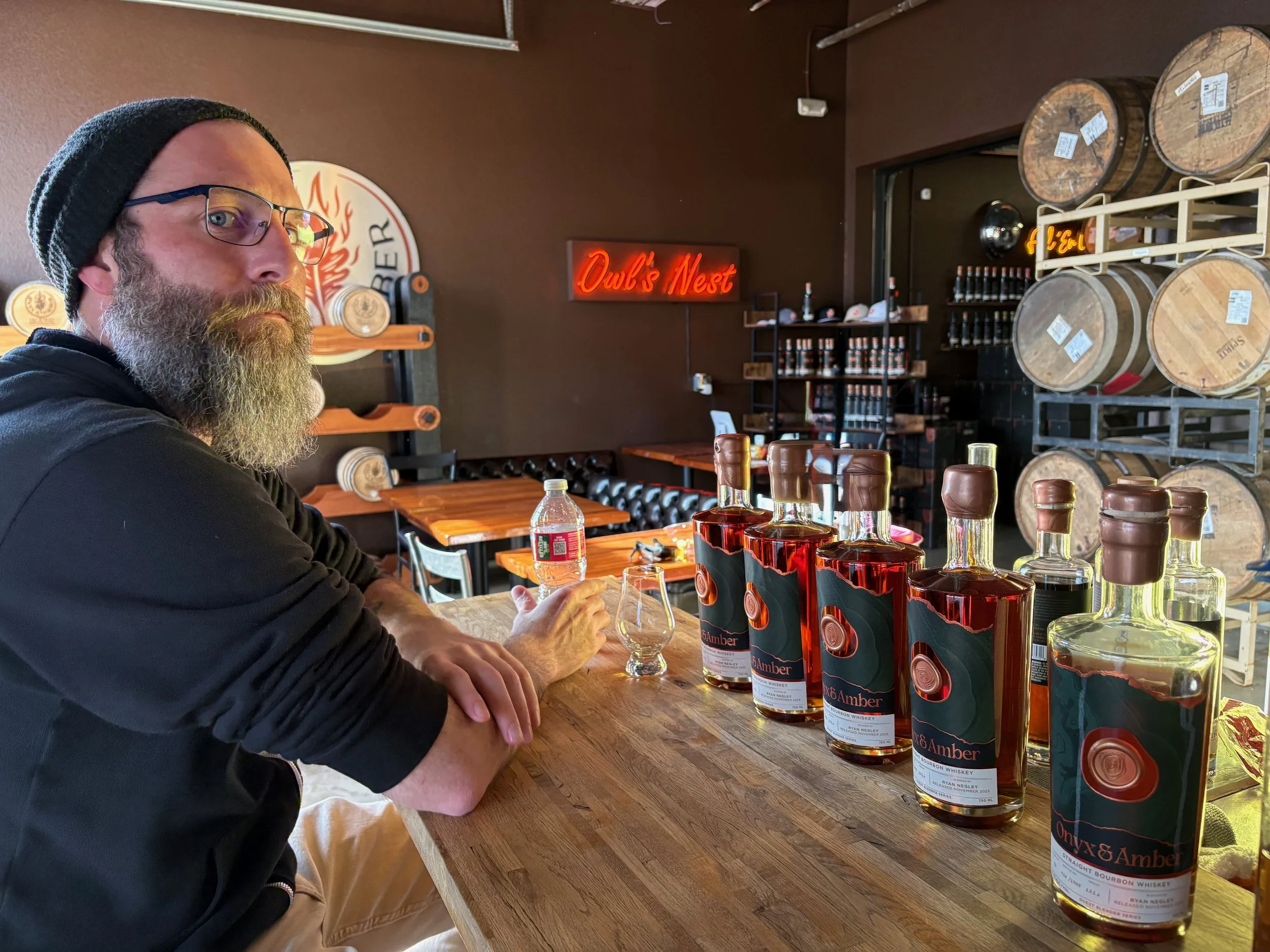 A man with a beard and glasses, wearing a black beanie and black shirt, is sitting at a wooden bar counter in a whiskey distillery or tasting room. Several bottles of whiskey are lined up on the bar in front of him, with other bottles and barrels visible in the background.