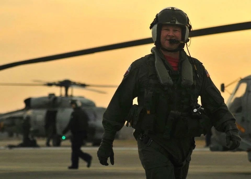 A military personnel in uniform walking on an airfield with helicopters in the background during sunset, wearing a helmet with a headset.