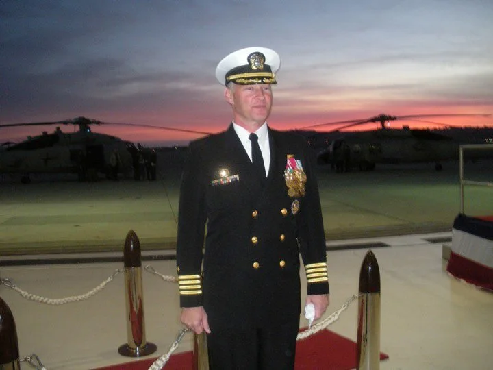 A military officer in formal uniform with medals standing on a red carpet at an airfield during sunset with helicopters in the background.