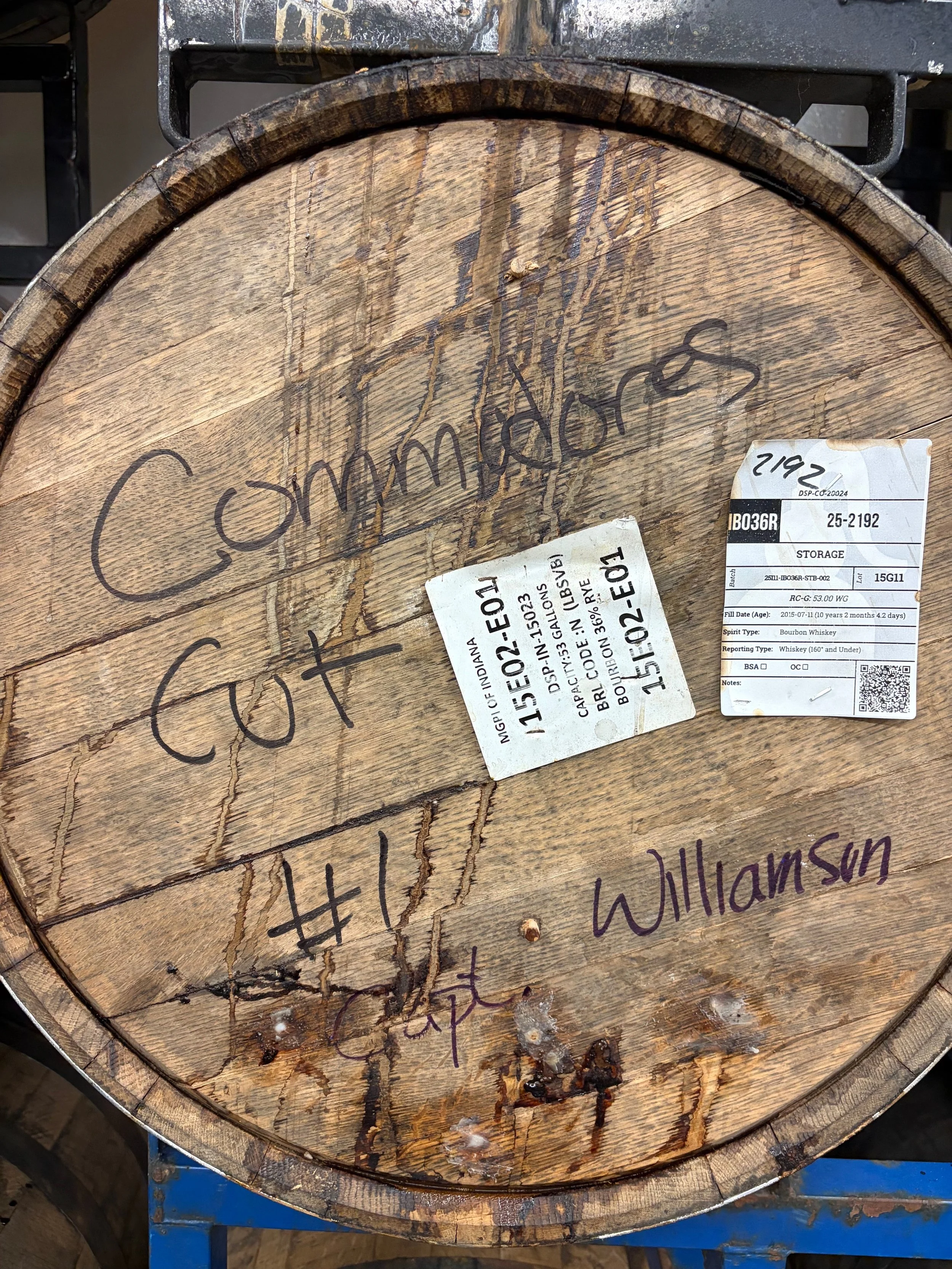 Close-up of a wooden barrel with handwritten markings and labels, including a barcode and other printed information, placed on a blue stand.