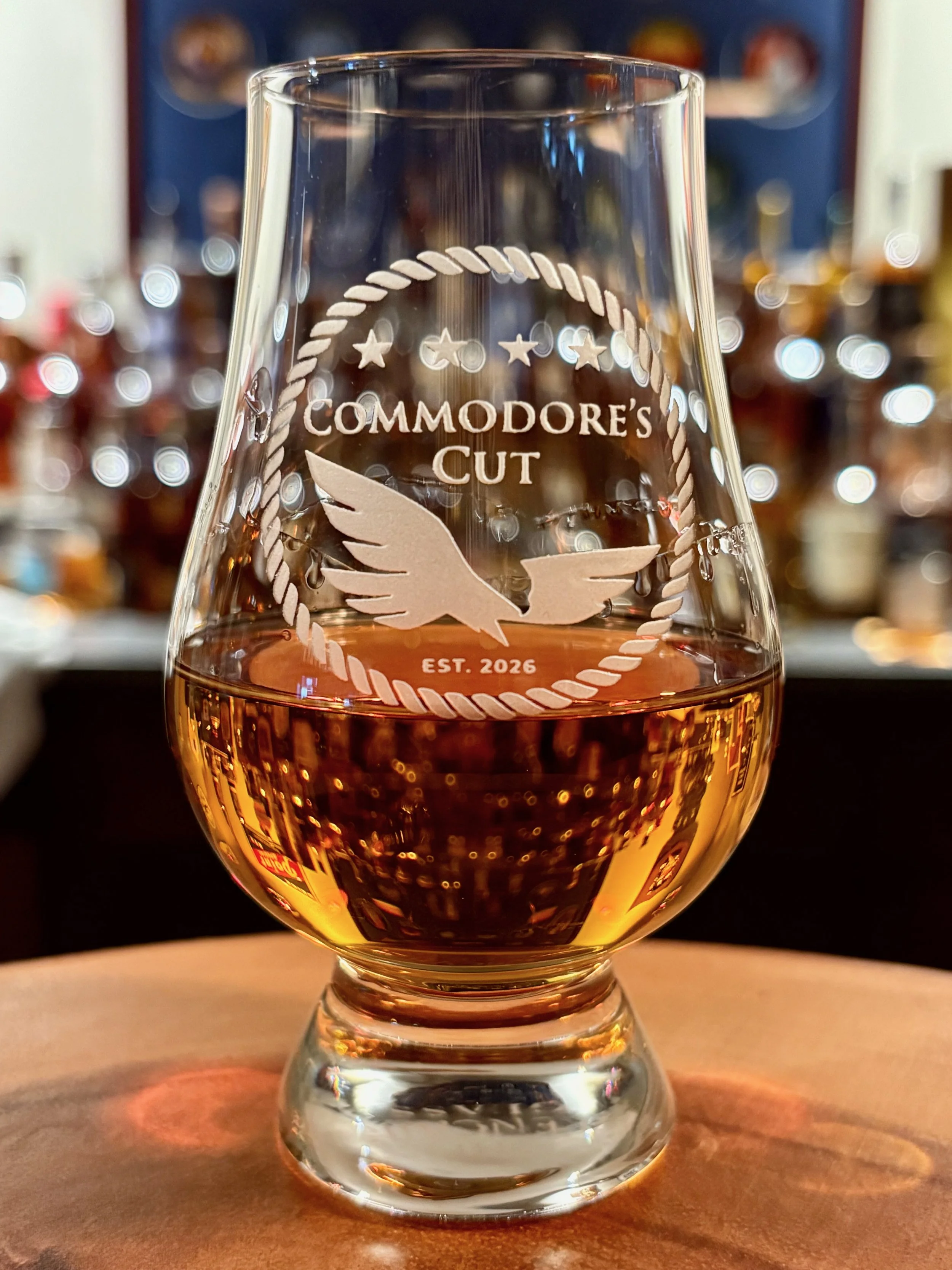 Close-up of a whiskey glass with the inscription 'Commodore's Cut, EST. 2026' and an engraved eagle logo, filled with a golden-colored beverage, on a wooden surface with blurred bar bottles in the background.