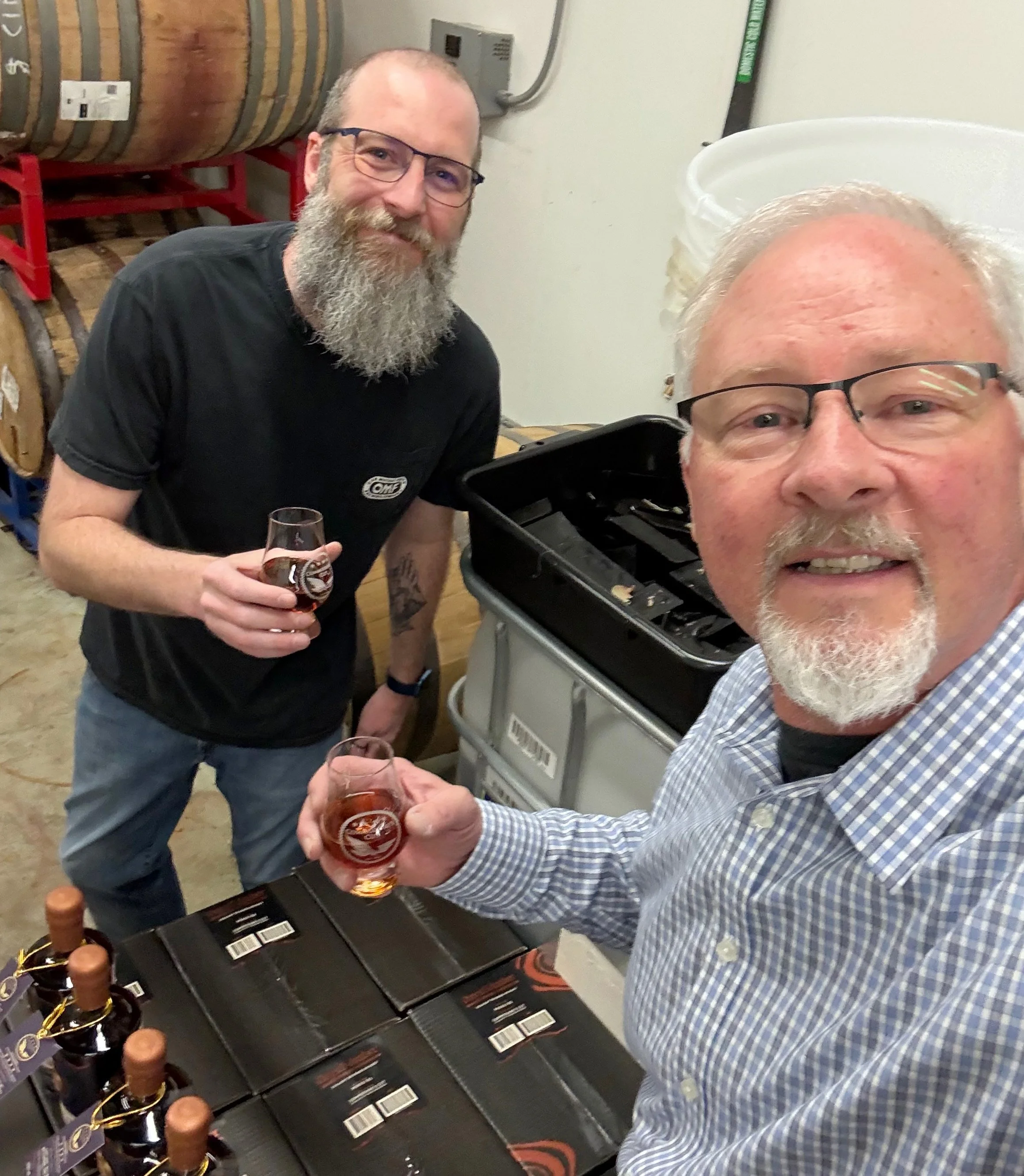 Two men, holding glasses of whiskey. One man has a gray beard and glasses, and the other man has a light-colored beard and glasses. There are bottles on the table in front of them, and behind them, there are whiskey barrels.