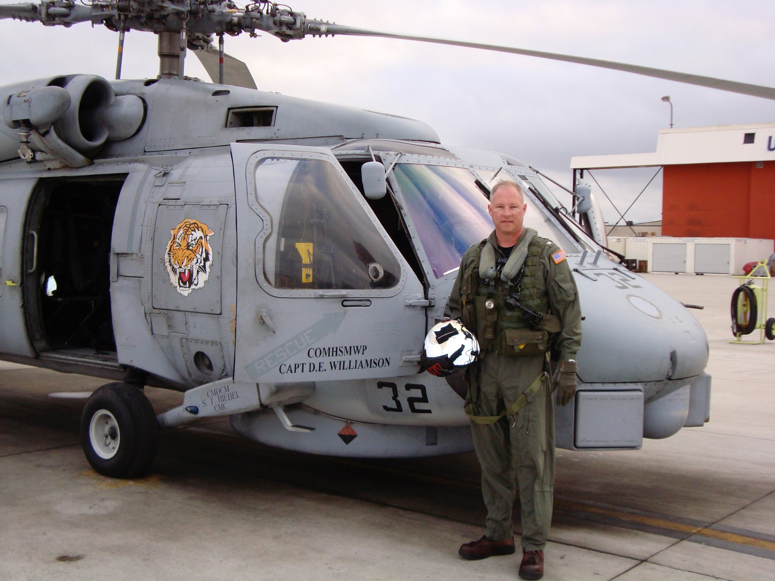 A military helicopter with a tiger emblem and the text 'COMMSMWP CAPT D.E. WILLIAMSON' on its side. A man in military uniform stands beside it, holding a helmet.