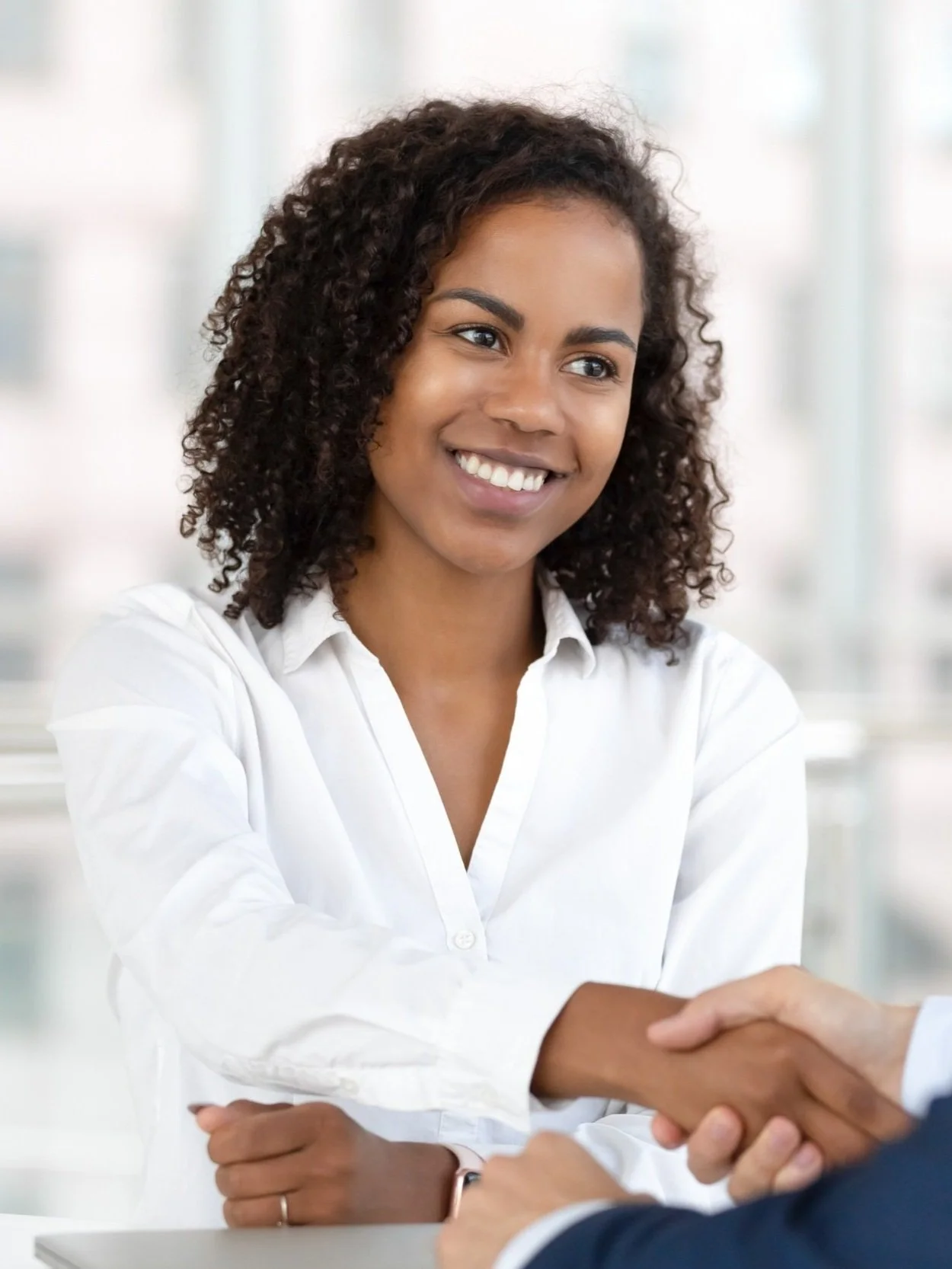 A smiling woman with curly hair, wearing a white shirt, shaking hands with a person in a business setting.