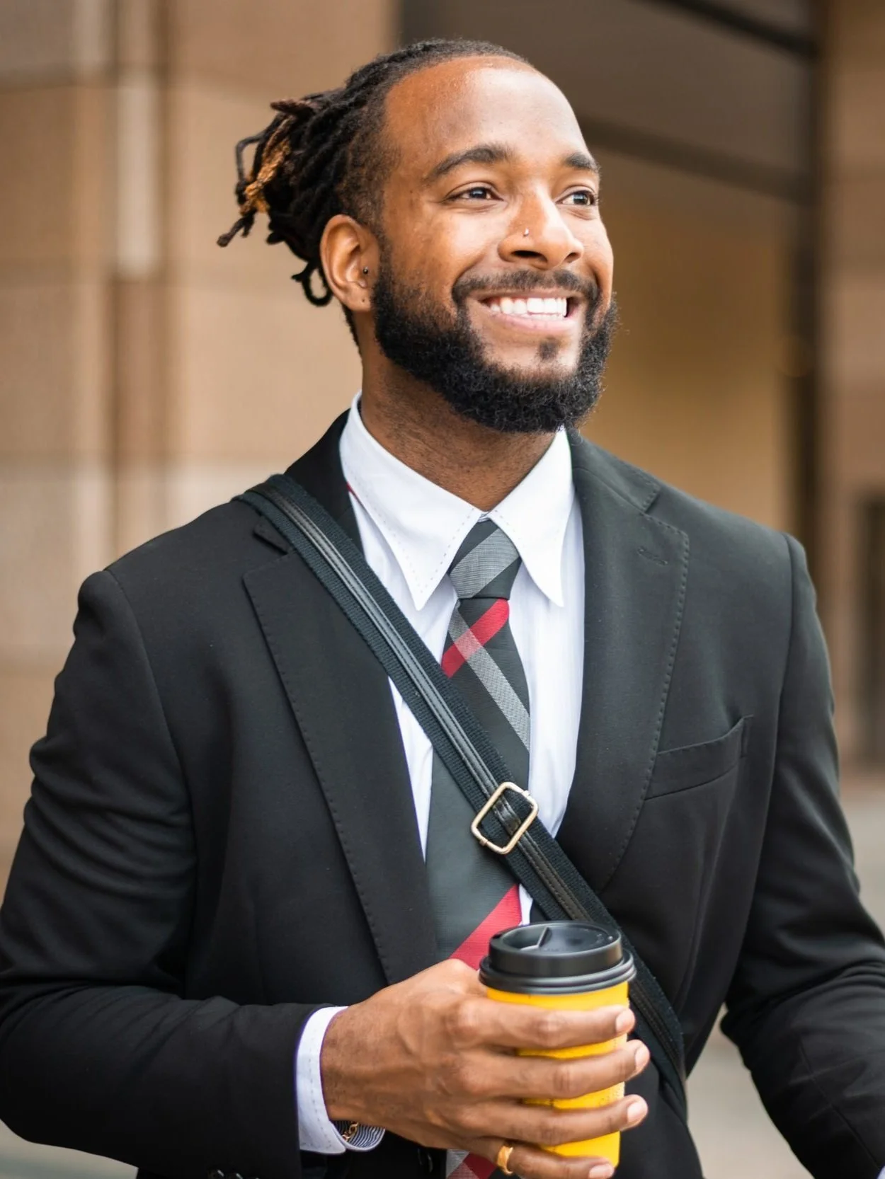 A smiling man with dreadlocks and a beard, wearing a black suit, or shirt, a white shirt, and a striped tie, holding a yellow disposable coffee cup with a black lid, standing outdoors.
