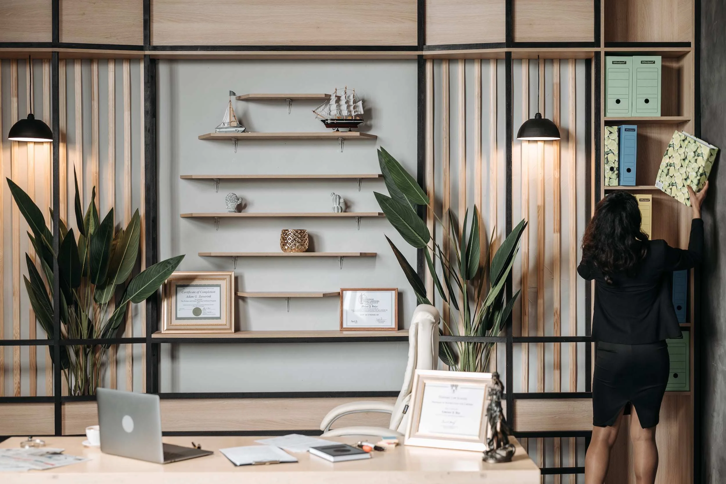 An office with a desk, laptop, and framed certificates. A woman with dark hair in a black dress places folders on a bookshelf, decorated with plants, shelves, and model ships.