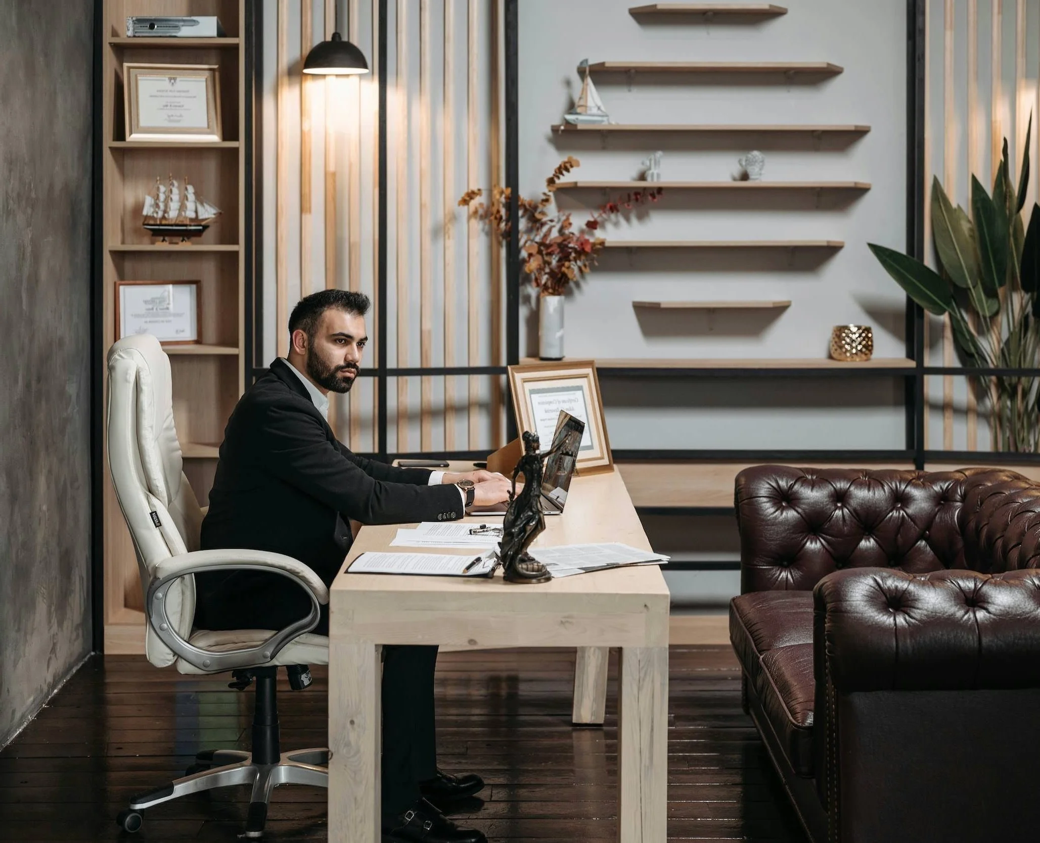 A man in business attire is sitting at a wooden desk with papers, a framed certificate, and a classical statue. He is working on a laptop in a modern office with wooden shelves, decorative items, and a leather sofa.