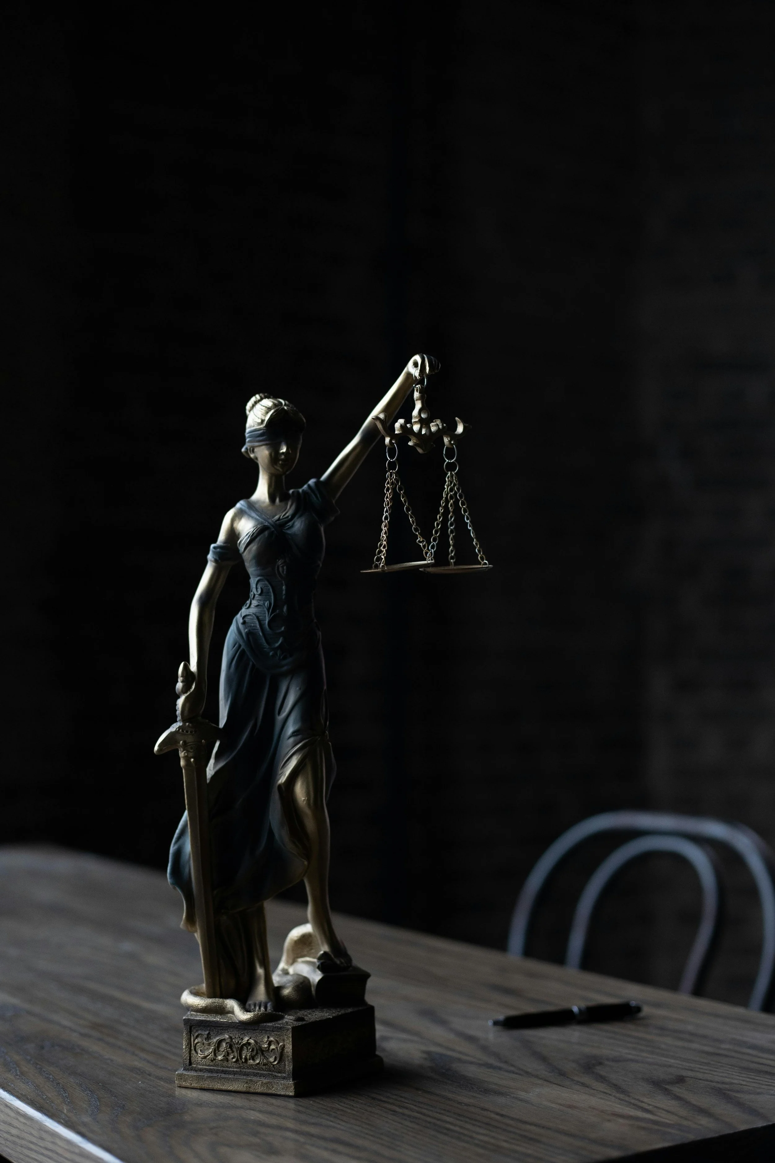 Statue of Lady Justice with a blindfold, holding scales in her right hand and a sword in her left, placed on a wooden table in a dimly lit room.